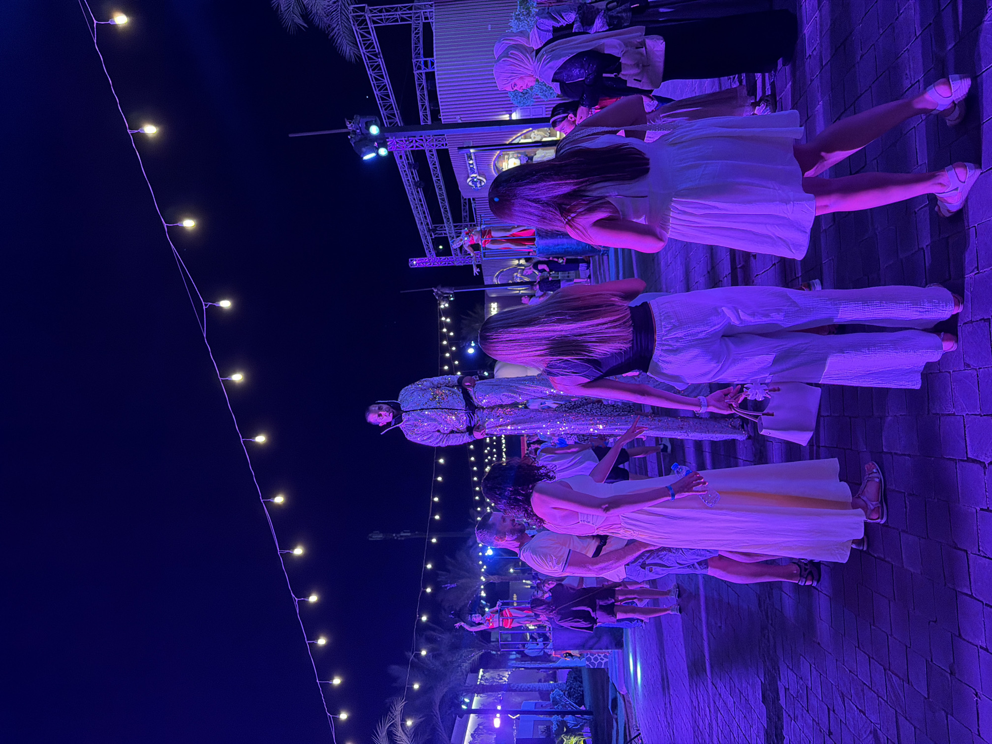 The image captures an outdoor evening gathering, likely a performance or celebration, in Sharm El Sheikh, Egypt. The scene is illuminated by vibrant purple stage lighting and a string of warm, yellow fairy lights draped across the frame. In the foreground, several individuals are visible, some facing away from the camera, dressed in casual evening wear. One person in the center stands out, dressed in a sequined, full-body outfit and appears to be a performer. Further back, a platform or stage is set up, where other individuals, including one in a red outfit, are also present, suggesting a planned event. The ground is paved with cobblestones, and palm trees are visible in the background, hinting at a resort or entertainment venue. The time of day is clearly night, given the dark sky and artificial lighting. The overall atmosphere appears festive and energetic. No visible text is present in the image.