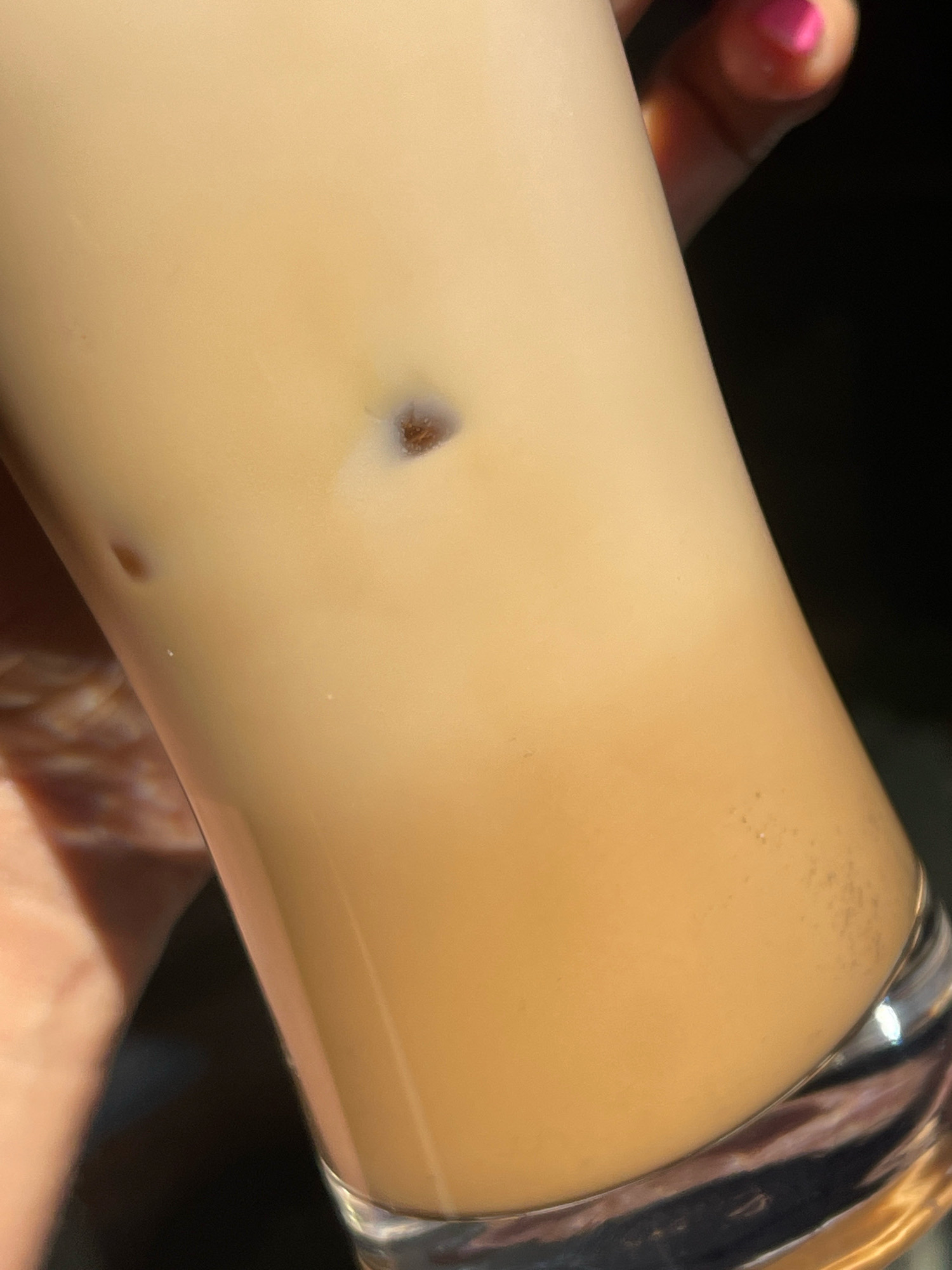 An extreme close-up shot focuses on a tall, clear glass containing a light brown beverage, likely coffee with milk or cream, and ice cubes. One ice cube is clearly visible slightly above the center of the frame, with a dark, irregular speck embedded within it. Another smaller, oval-shaped speck is visible to the left of the ice cube, floating in the liquid. The glass itself has a reflective quality, showing distortions of the surrounding environment. At the top right, a portion of a hand is visible, with a bright pink fingernail polish on the index finger. The background is dark and out of focus, suggesting an indoor setting or evening light. The image does not contain any elements that would place it in Karlovo, Bulgaria.