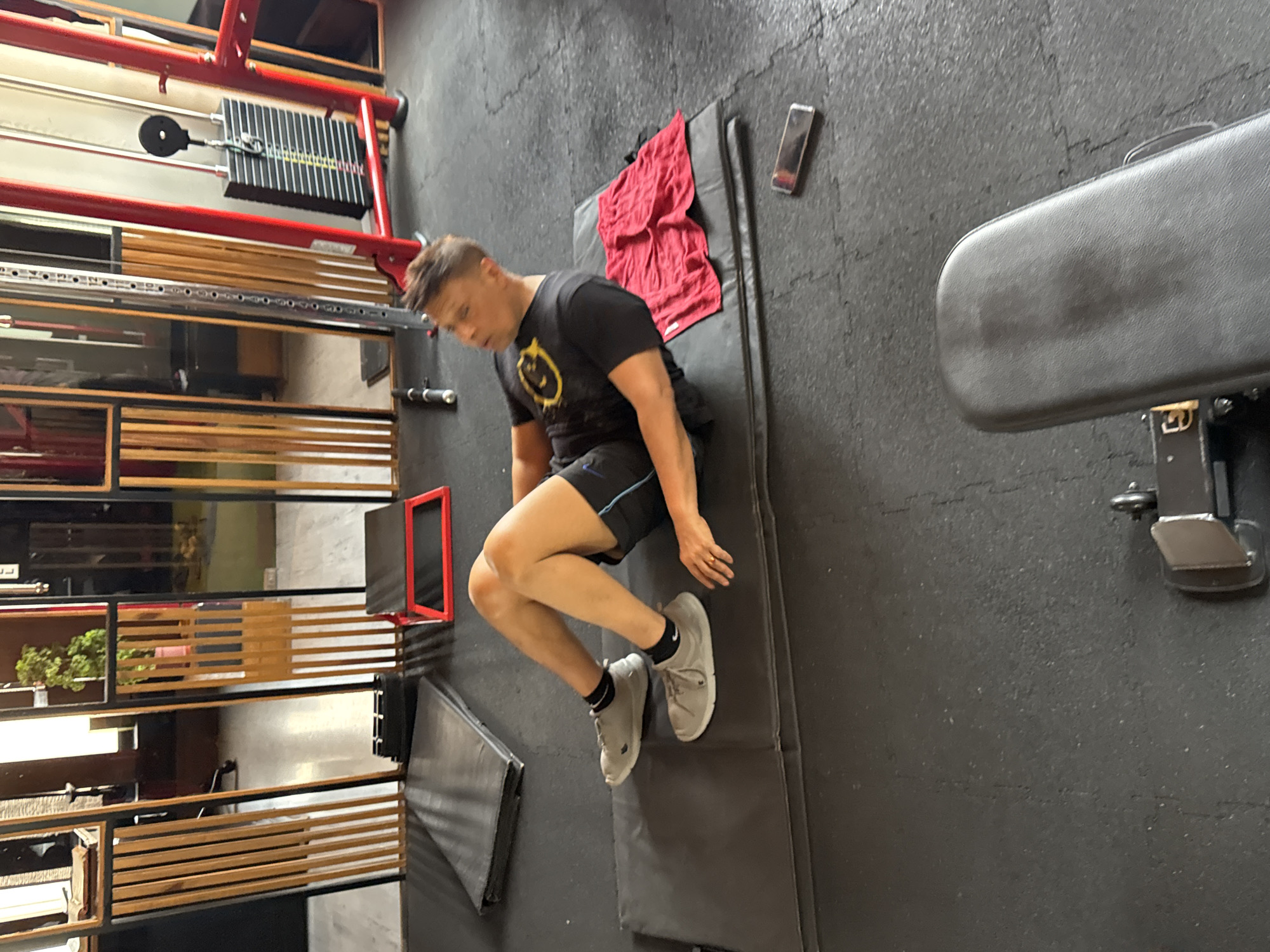 A man in a black t-shirt and shorts is performing a floor exercise on a gray mat in a gym. A red towel and a cell phone are near the top of the mat. A weight machine and shelving are to the left of the mat. A weight bench is to the right of the mat. The gym has gray flooring. The location cannot be determined to be Kolkata, India from the image.