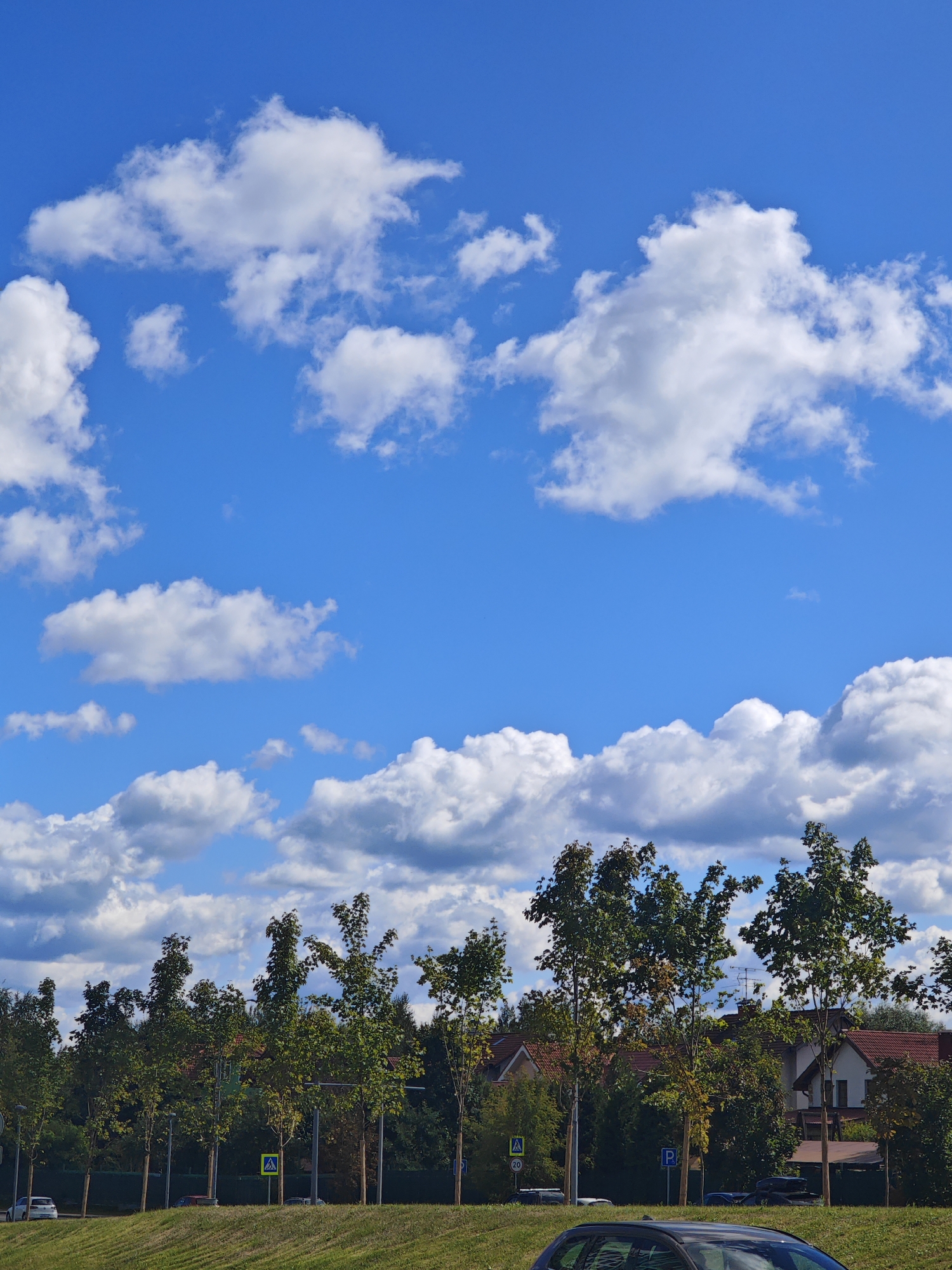 The image depicts a residential street in Moscow, Russia, under a bright blue sky filled with fluffy white clouds. The foreground shows a neatly mown grassy slope, leading to a street lined with young, deciduous trees. A white car is parked on the left side of the street. Further down the street, several houses with red-tiled roofs are visible, interspersed with dense green foliage. Traffic signs, including a pedestrian crossing sign and a parking sign, are present along the street. The scene conveys a peaceful, suburban atmosphere typical of a summer day in Moscow.