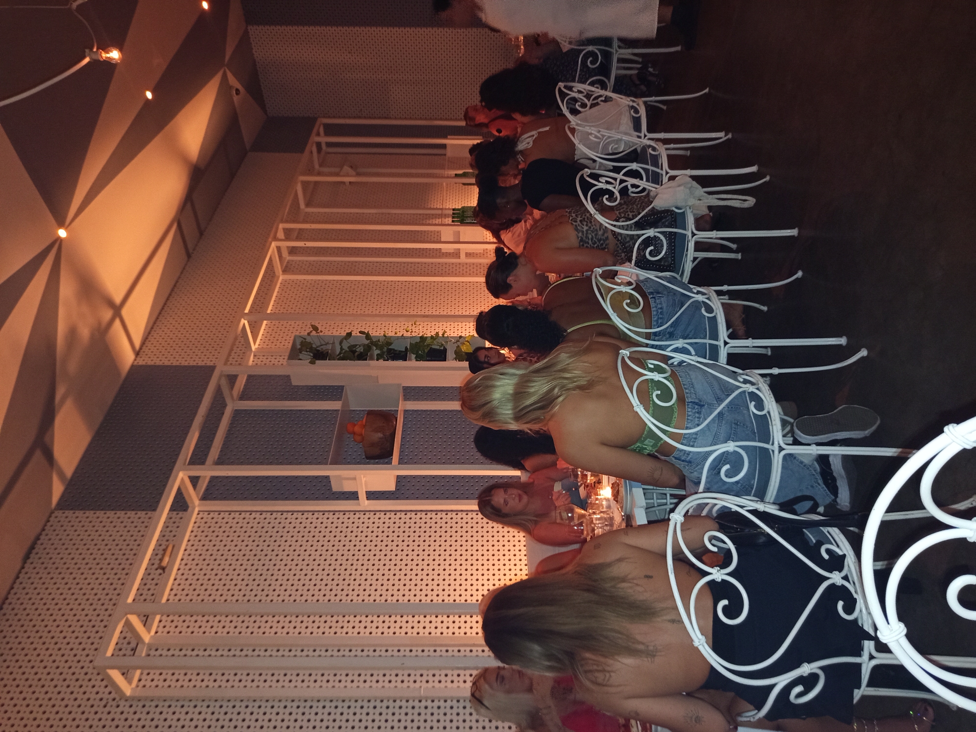 The image shows a long dining table in what appears to be a restaurant setting in North Kuta, Indonesia. A group of people, mainly women, are seated at the table. The restaurant features a white-lattice wall design with built-in shelves holding plants and decorative objects. The chairs are white with ornate, scrolled backs. The light is dim, suggesting an evening setting. A mix of clothing styles is visible.