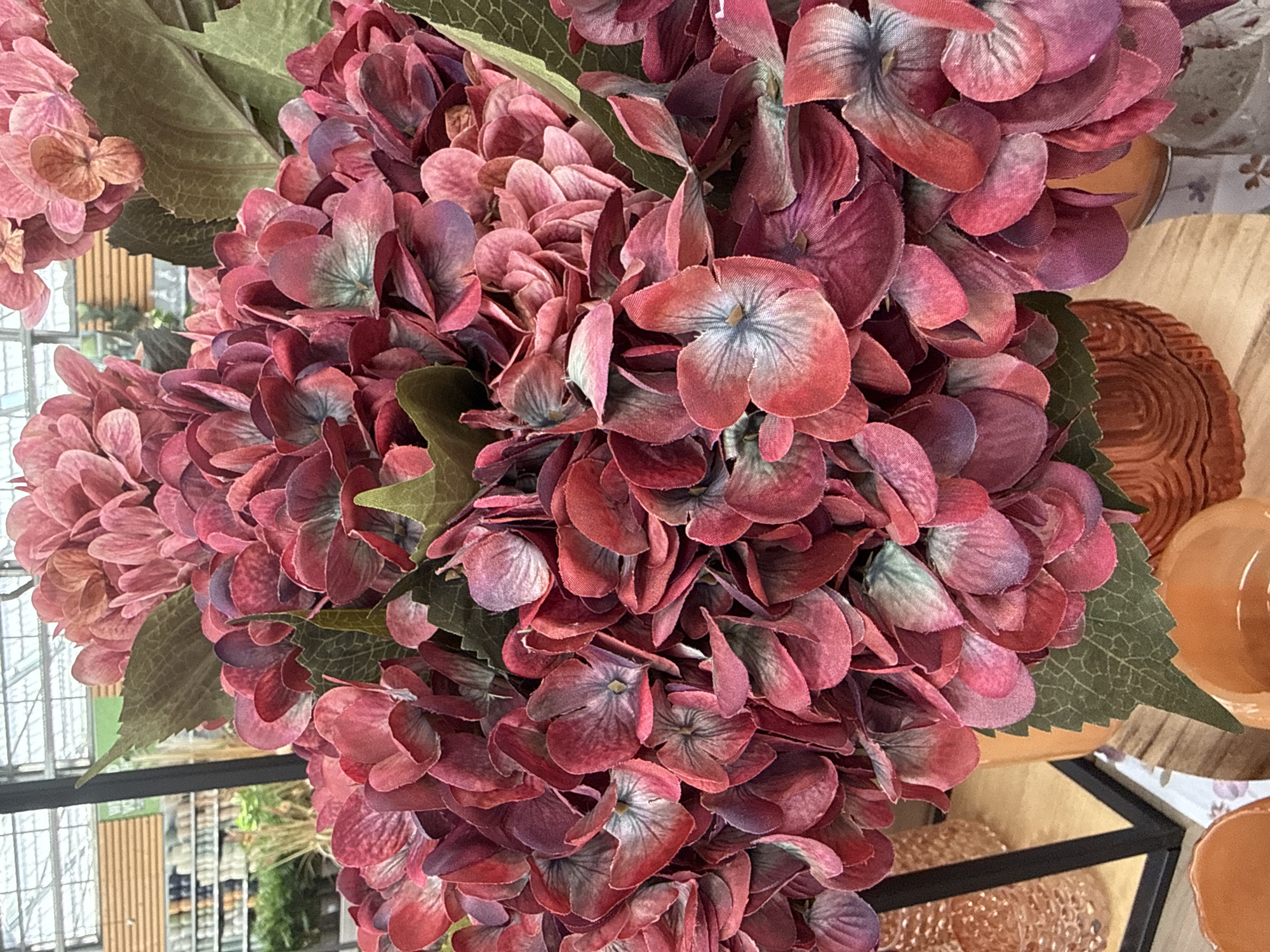 A close-up, eye-level shot captures a large bouquet of artificial hydrangeas in shades of deep pink, crimson, and muted purple. The flowers are densely packed, with their textured petals overlapping and creating a sense of volume. Some of the individual blooms reveal a subtle gradient of color, transitioning from a lighter pink at the edges to a darker hue towards the center, where faint blue veining is visible. Interspersed among the flowers are several realistic-looking green hydrangea leaves with visible vein patterns. The bouquet is arranged in an orange, textured ceramic vase, partially visible at the bottom center of the frame. To the left, a black metal frame, likely part of a display shelf, encloses a glimpse of a store interior. Rows of merchandise, possibly gardening supplies or home decor, are visible in the blurred background, suggesting the setting is a retail environment. The overall lighting is bright, characteristic of an indoor commercial space. There are no people or animals present in the image. The context of Fritzlar, Germany is not evident from the visual information within the photograph.