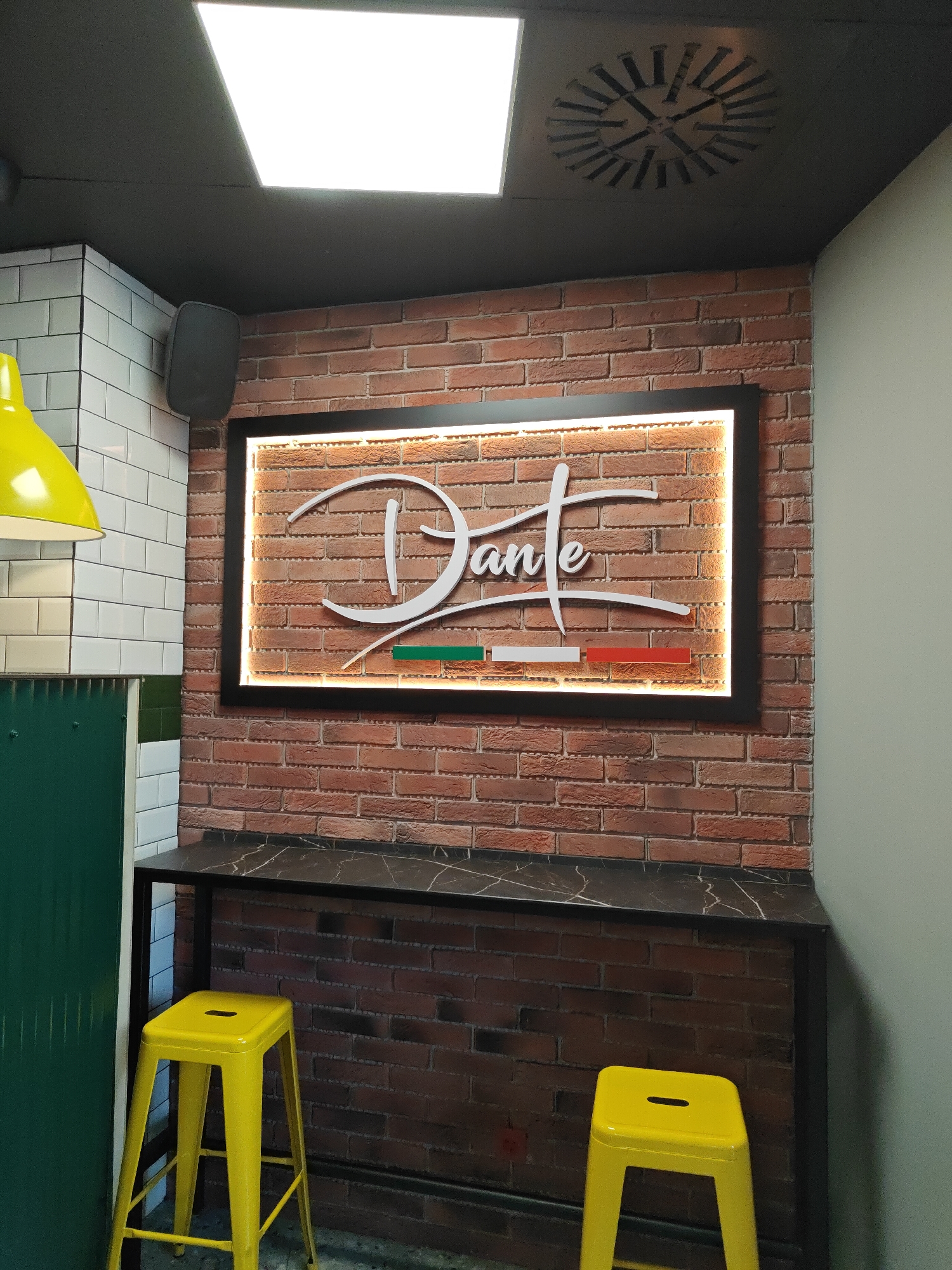 The image captures an interior scene featuring a "Dante" branded restaurant. The backdrop consists of a red brick wall on which a rectangular, black-framed light sign displaying the restaurant's name in cursive is mounted. The colors of the Italian flag are also featured in the sign. Below the sign, a black countertop table is situated. Two yellow stools are positioned in front of the table. To the left of the sign is a white-tiled wall with a section of green tile. A yellow lamp is visible above the tiles. A speaker is affixed to the wall above the counter. The ceiling is dark gray and features a square light fixture and a circular ventilation grille. The right side of the scene shows a light gray wall. The city and country in which the scene is located, Sofia, Bulgaria, cannot be confirmed solely from this image.