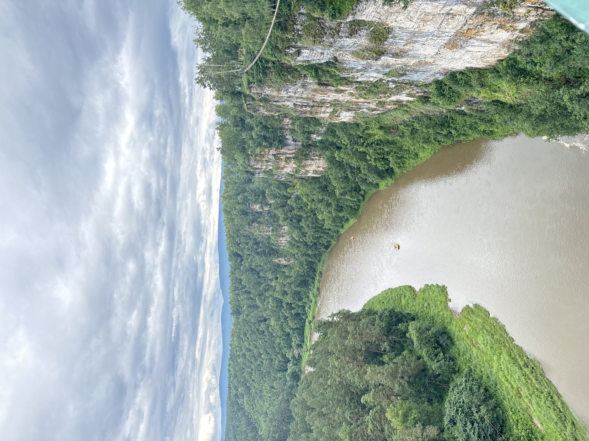 The image depicts a natural landscape with a river flowing through a densely forested gorge. The viewpoint is from a high vantage point, looking down and across the scene. The cliff face on the right is rocky and covered in green vegetation. A suspension bridge stretches across the gorge from the cliff, with a yellow marker visible on the bridge. The river below is a murky brown and has two small yellow objects, possibly kayaks or rafts, on its surface. The surrounding area is covered in lush green trees, with some patches of grass on the riverbank. The sky is filled with dramatic, overcast clouds, suggesting a cloudy or stormy day. The lighting indicates it is daytime, but the overcast conditions reduce direct sunlight. The overall mood is one of natural grandeur and perhaps a sense of adventure, given the presence of the bridge and watercraft. The location context provided is Kul'metovo, Russia, which aligns with a rugged, forested natural environment. There is no visible text in the image.