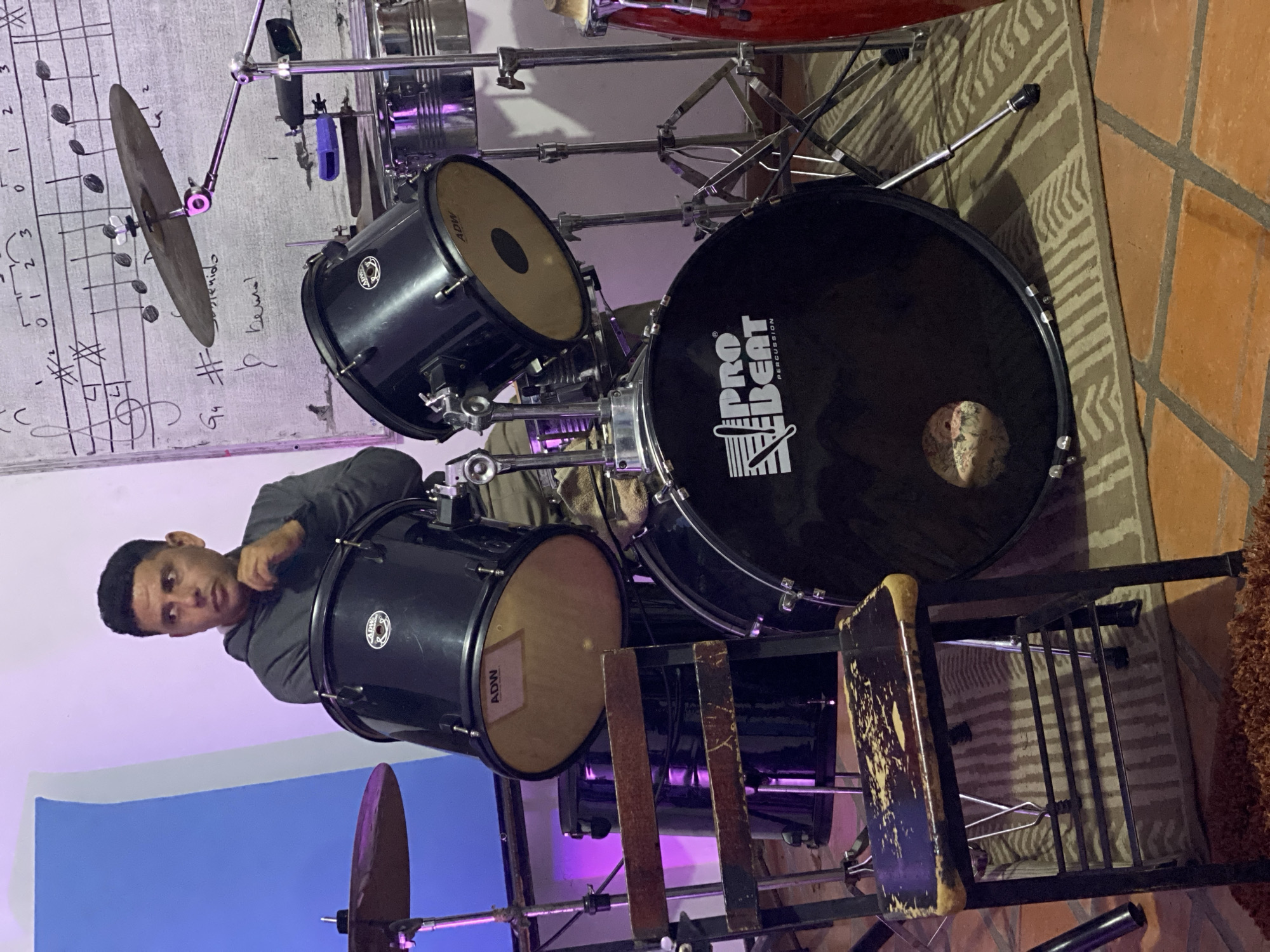 A man stands behind a black drum set in a room, presumably in Tinaquillo, Venezuela. The drum set consists of a bass drum with a "Pro Beat Percussion" logo, two smaller drums, and cymbals. The man is wearing a grey long-sleeved shirt. Sheet music hangs on the wall behind the drum set. A worn chair is visible near the drums. The room is lit with both natural and artificial lighting, including a blue-purple light source. The floor is a mix of tile and rug.