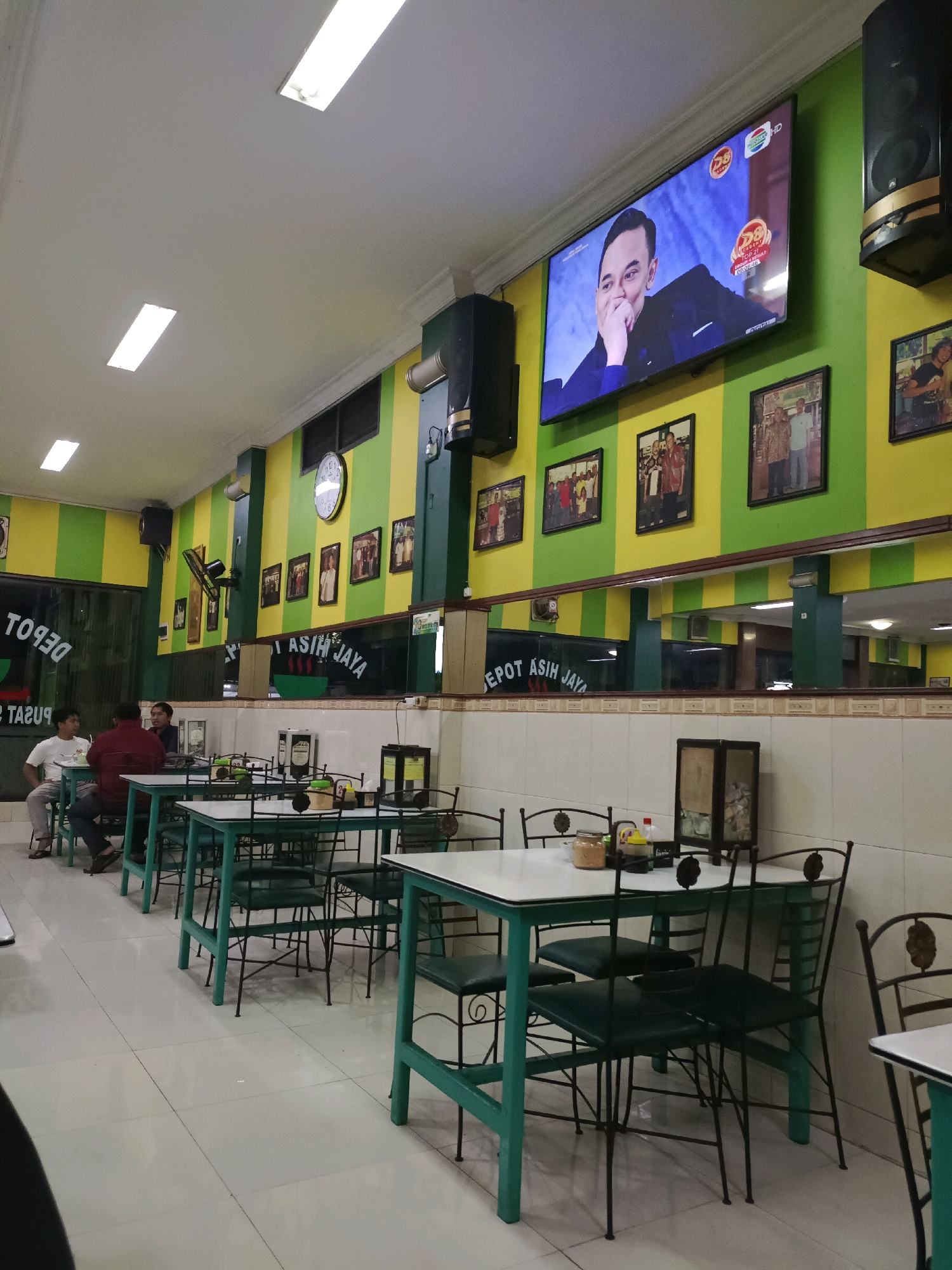 The image depicts the interior of a restaurant or "Depot" in Dapur, Indonesia. The walls are painted with alternating vertical stripes of bright green and yellow. A large flat-screen television is mounted on the wall, displaying a man in a suit. Several framed photographs are also displayed on the walls. The restaurant features numerous tables with green metal frames and white tabletops, surrounded by black metal chairs with green seats. Some tables are set with condiments and small decorative items. In the foreground, three men are seated at a table on the left side of the image, engaged in conversation. The floor is tiled in a light cream color. The overall atmosphere appears casual and communal.