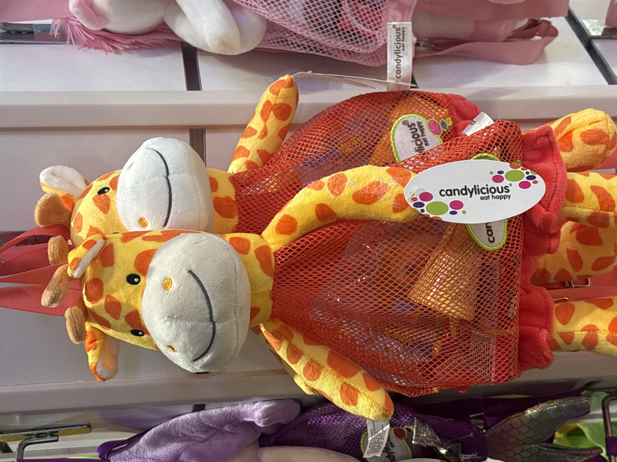 In Dubai, United Arab Emirates, a close-up shot captures two plush giraffe toys hanging in a store. Both giraffes are yellow with orange spots and wear red mesh bags around their lower bodies, revealing some contents within. The giraffe in the foreground is more prominently featured, with its head and upper body visible. A white oval tag with colorful polka dots and the text "candylilicious eat happy" is attached to its mesh bag. To the right, another giraffe is partially visible, also wearing a red mesh bag. The background consists of a white wall and other merchandise, including a purple bag with a scale-like texture to the left and a pink plush toy with pink hair to the far right. The lighting suggests an indoor retail environment.