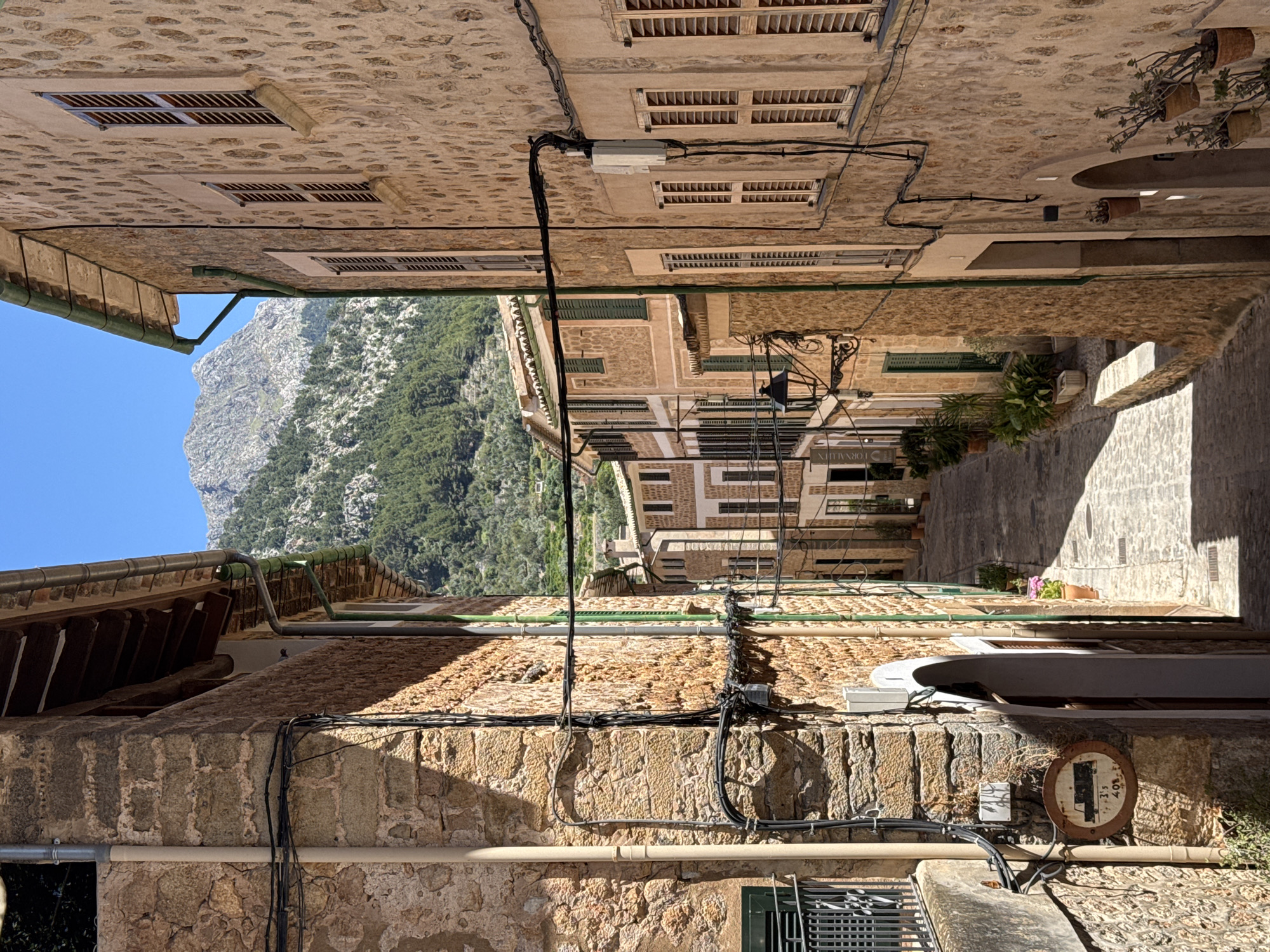 The image captures a narrow street view in Fornalutx, Spain. Stone buildings line both sides of the street, typical of the town's architecture. Mountains rise prominently in the background, visible at the end of the street. Electrical wires and pipes run along the facades of the buildings. Shutters cover some of the windows. A round sign is affixed to one of the walls. The lighting is uneven, with some areas in direct sunlight and others in shadow. No people are visible in the frame.