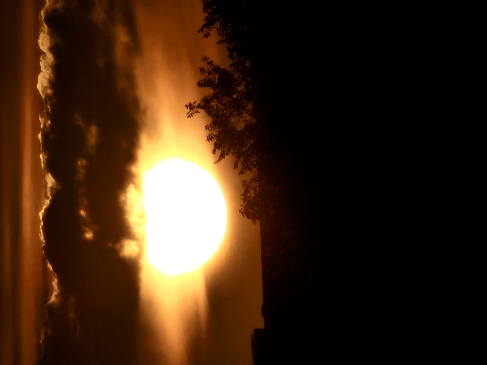 The image depicts a dramatic sunset with the sun partially obscured by dark clouds. The sun appears as a bright, glowing orb in the upper center of the frame, casting an orange and yellow hue across the sky. Silhouetted against the vibrant sky are the dark shapes of trees and what appears to be a building on the left. The lower portion of the image is a solid black silhouette, suggesting a foreground element like a hill or the ground. The scene evokes a sense of twilight in the city of Khimki, Russia, characterized by its natural beauty and urban landscape.