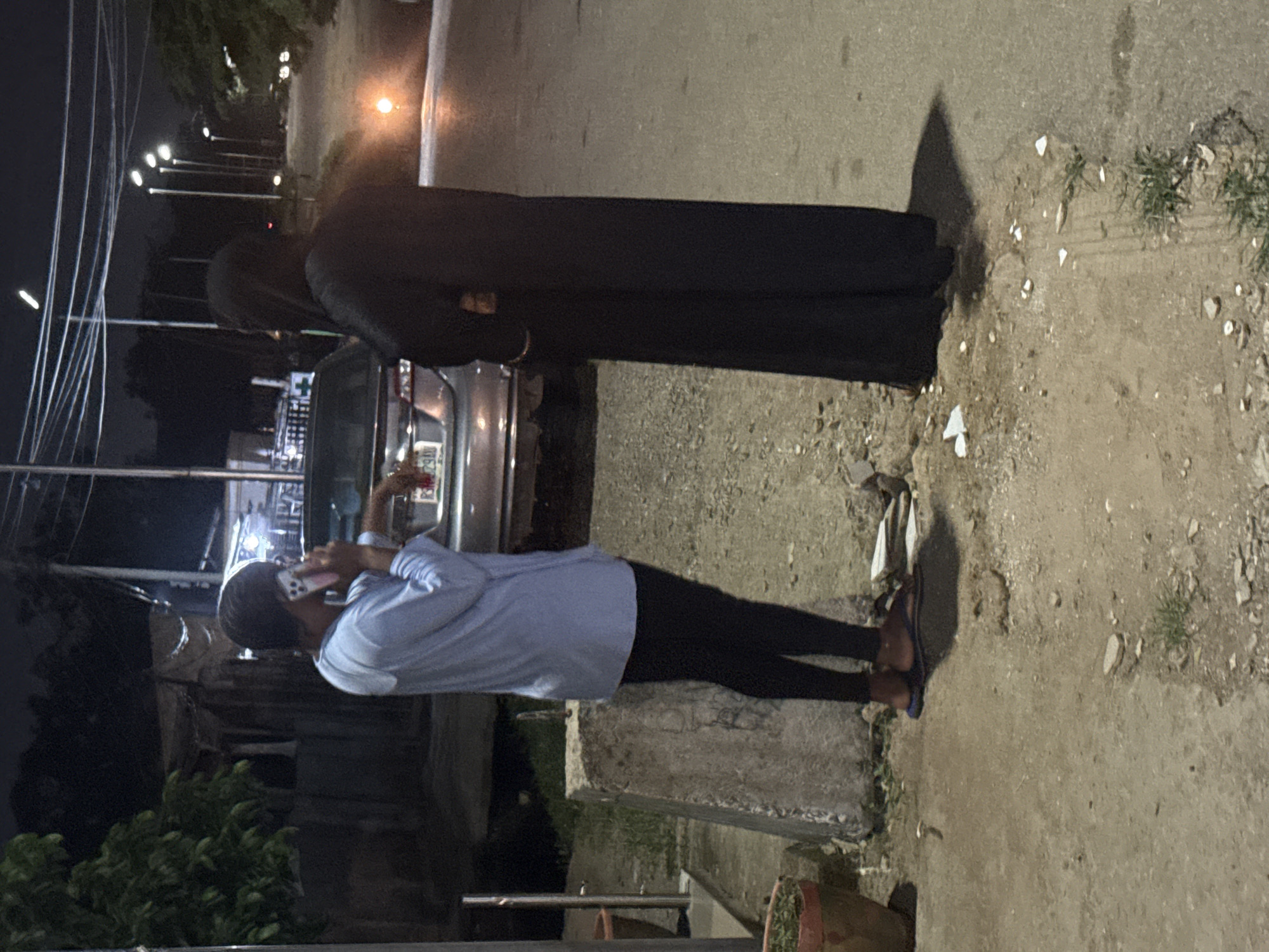 In Abuja, Nigeria, at night, two individuals stand near a parked vehicle on a dirt and asphalt road. One person, wearing a blue long-sleeved shirt and black pants, is on their phone, leaning on the car. The second individual, draped in a black abaya, stands facing away from the viewer, looking towards a street with multiple lampposts illuminating the scene. In the background, there are structures with tarpaulin roofs, and the overall environment suggests an urban setting with some signs of informal development.