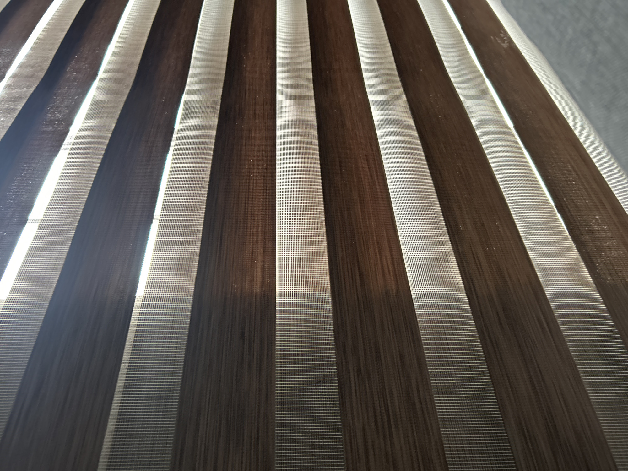 The image shows a close-up view of striped window blinds. The stripes alternate between a dark brown/black color and a light, possibly white or beige, color. The blinds appear to be made of a fabric-like material with a fine mesh texture. Light is visible shining through the lighter stripes. The scene does not provide information about a specific location or city.