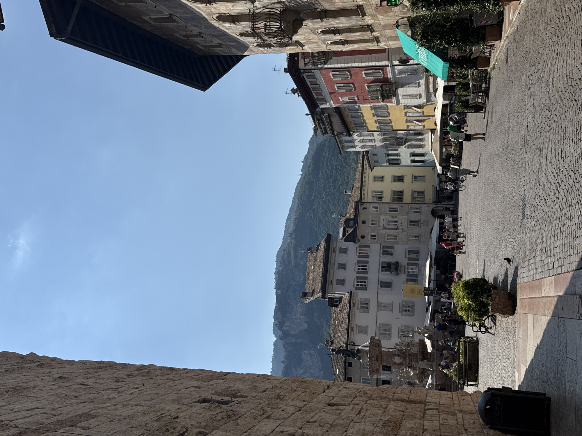The image depicts Piazza Duomo in Trento, Italy, on a clear day. The square is paved with cobblestones, and on the right side, a building with arched windows and balconies is visible, its facade showing signs of age. To the left, a tall stone wall occupies a significant portion of the frame.
In the background, a row of colorful buildings lines the square, including structures painted in yellow and red, with tiled roofs. A fountain featuring a statue of Neptune is situated in the mid-ground. Numerous people are spread across the square, some walking, others gathered near the fountain or seated at outdoor cafe tables under umbrellas. The presence of benches, planters with greenery, and a trash can suggests a public space designed for leisure and commerce.
A bird is seen walking on the cobblestones in the foreground. The environment is characteristic of an Italian city square, with a backdrop of hills or mountains under a bright blue sky. The overall atmosphere appears to be one of daytime activity and pleasant weather.