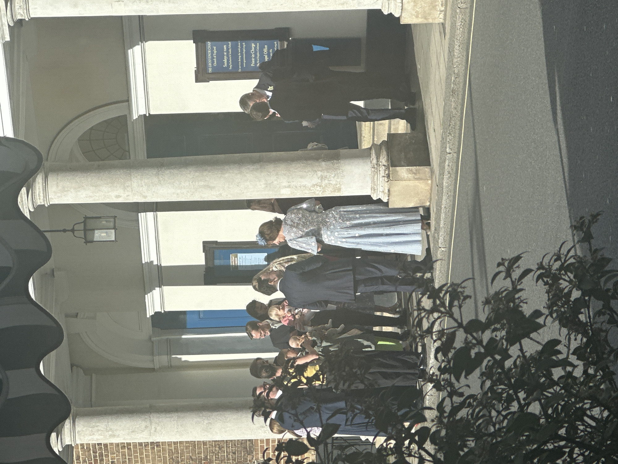 The image depicts the Grosvenor Chapel, a Church of England located in the city of London, United Kingdom. A group of individuals is positioned on the steps in front of the church entrance. The group includes men in suits and women wearing dresses; there is also at least one child being held. In the background, a sign visible on the building indicates service times and contact information for the chapel office. The setting is a sunny day, with shadows cast on the sidewalk. Some greenery is visible on the right side of the image. The architecture features columns and arched windows.