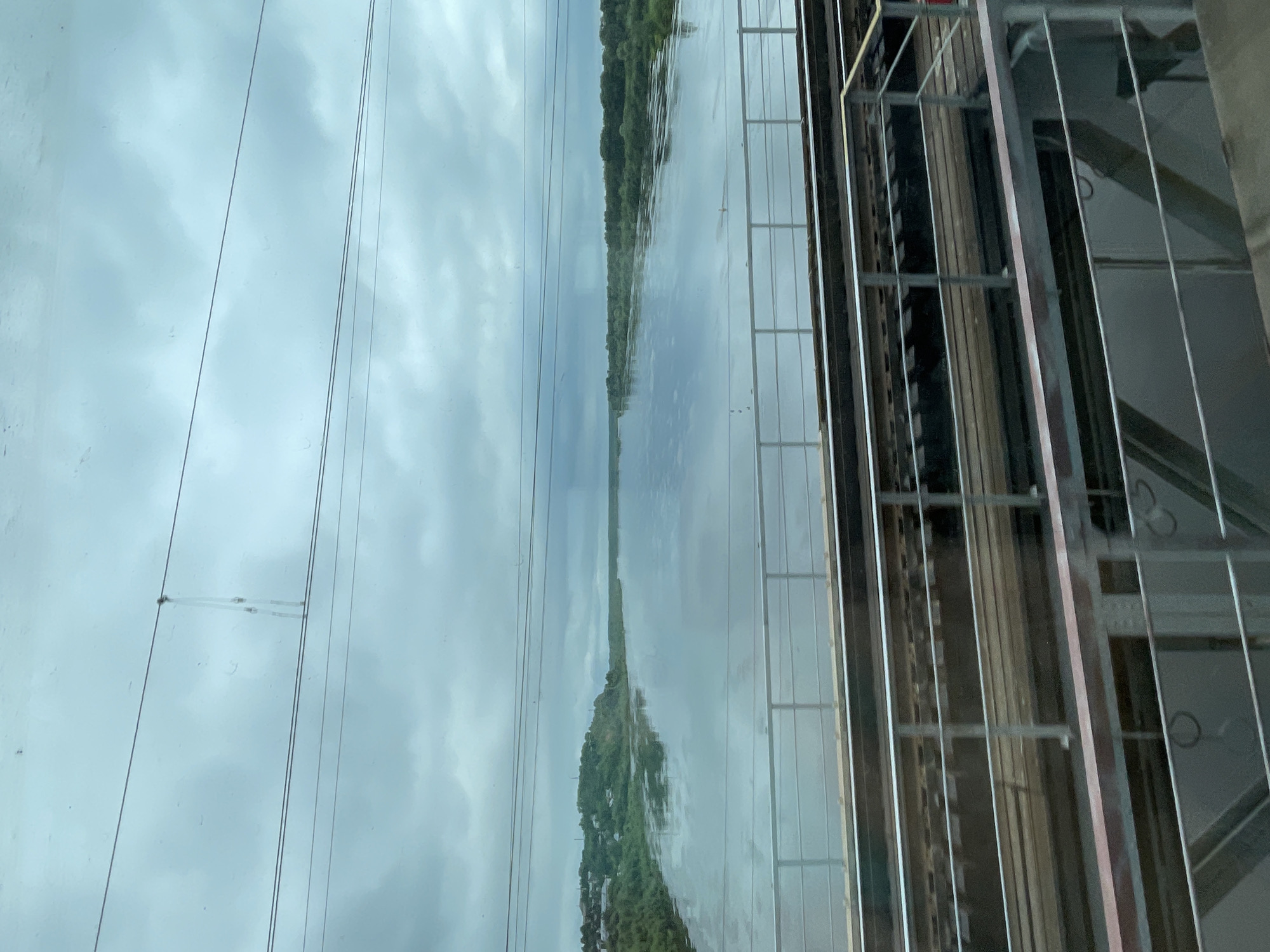 The image captures a view from a train, looking out over a wide river under an overcast sky. In the foreground, train tracks run horizontally across the frame, separated by a metal railing. The river occupies the midground and background, reflecting the cloudy sky and the lush green banks dotted with trees and some buildings on the left side. The scene appears to be in the city of Kotelnich, Russia, given the context provided. The overall atmosphere is calm and somewhat muted due to the cloudy weather.