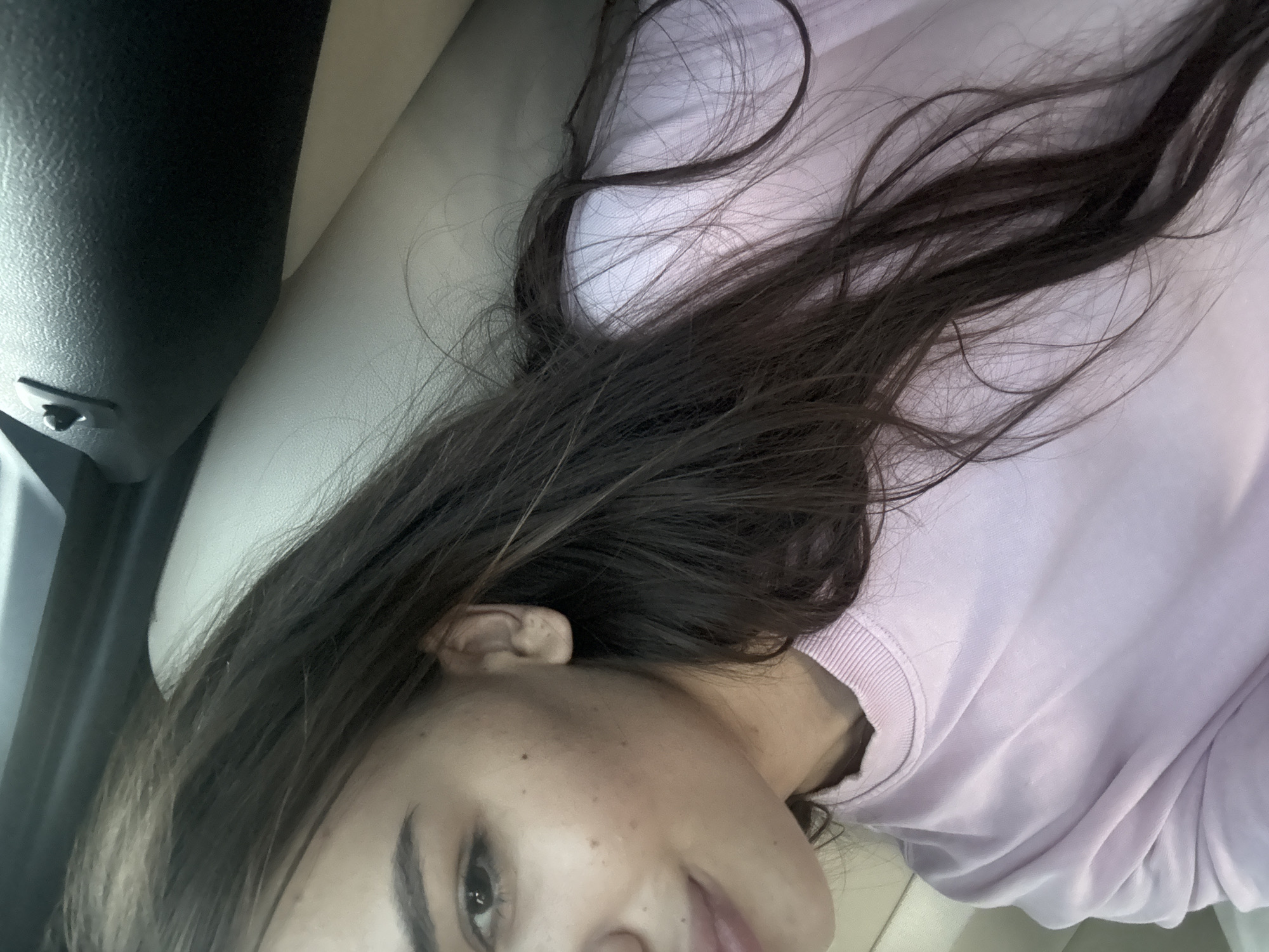 A medium close-up selfie shows a young woman with long, dark hair and fair skin. She is wearing a light pink sweatshirt and lying down. Her head is tilted to the side, and her hair is spread out. Her left ear is visible. The background appears to be the interior of a vehicle, with a light-colored seat and a dark dashboard. The image is taken in Sărata-Mereșeni, Moldova.