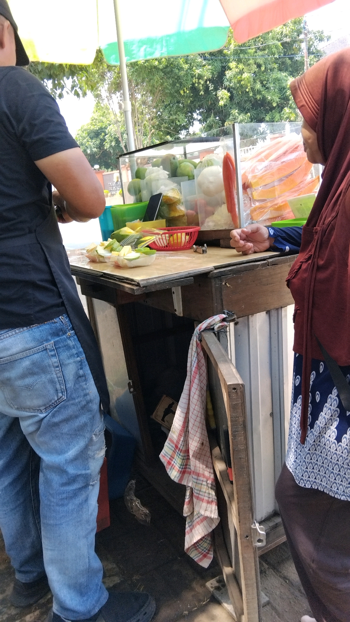 In the city of Batang, Indonesia, a street vendor prepares fruit for sale. The vendor, wearing a black t-shirt and blue jeans, stands behind a wooden cart. A woman in a maroon hijab and blue patterned tunic stands to the right. The cart displays various fruits, including green mangoes, white cabbage, and sliced papayas. The scene appears to be an outdoor market with trees and a tent providing shade.