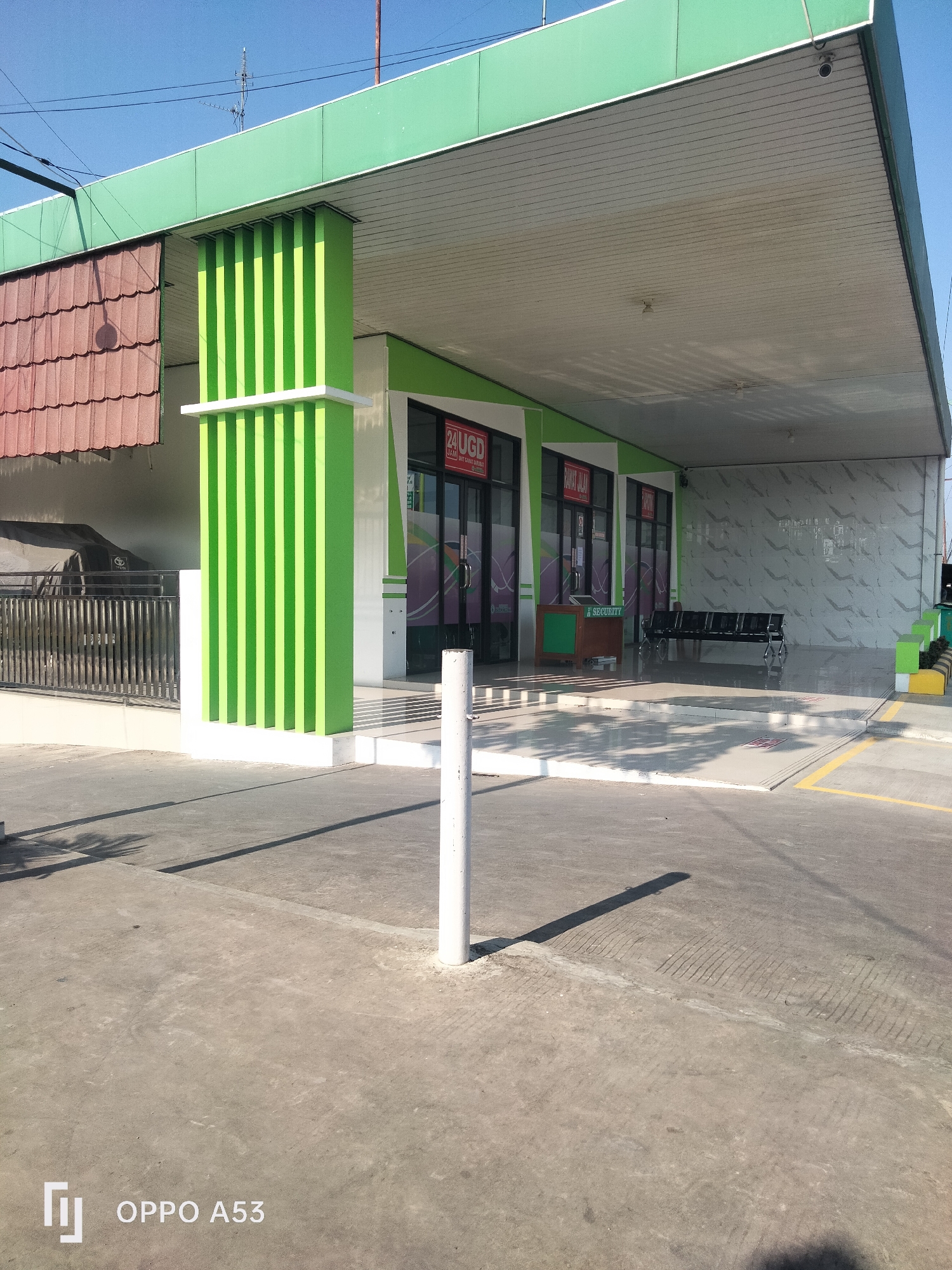 An outdoor, eye-level, full shot shows the entrance to a building with a green overhang supported by green, vertically striped columns in Karawang, Indonesia. The building's facade is predominantly white with green accents. To the left, a section of a building with a brown tiled roof is visible. A black metal fence is in front of a dark-colored vehicle parked under a cover. The foreground consists of a concrete surface with shadows cast by the building's overhang and a white pole.
The building's entrance features glass doors with signage. One sign reads "24 UGD" in red and white, indicating 24-hour emergency services. Another sign reads "RAWAT JALAN" in red and white, meaning outpatient care. Inside one of the entrances, a green counter with the word "SECURITY" is visible.
Further into the building's entrance area, a row of black plastic chairs is arranged against a wall with a textured, marble-like finish. The area is paved with light-colored tiles. The sky above is clear and blue. The overall impression is of a clean, modern medical facility or administrative building. The presence of the "OPPO A53" watermark suggests the image was captured with that model of smartphone.