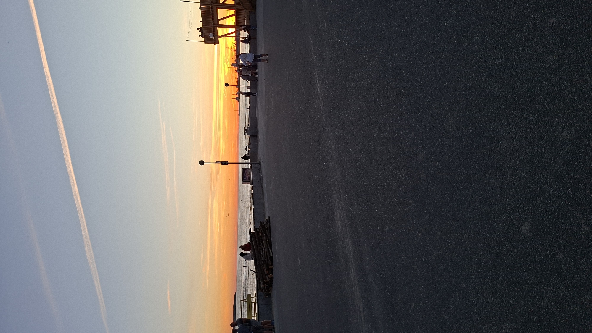 The image depicts a sunset scene at the Tallinn City Hall Square, also known as Viru väljak, in Tallinn, Estonia. A group of people are visible standing on a paved area, with the sea and a vibrant sunset in the background. The sky is illuminated with warm orange and yellow hues, indicating the sun is setting. In the distance, there are hints of industrial structures and ships along the coastline. A wooden structure with lights strung across it is present on the right side of the image. Several individuals are casually dressed and appear to be enjoying the evening. The foreground is dominated by the dark asphalt of the square, with a pile of logs to the left, suggesting a possible event or preparation for one. A yellow bench is situated near the edge of the paved area, facing the sea.