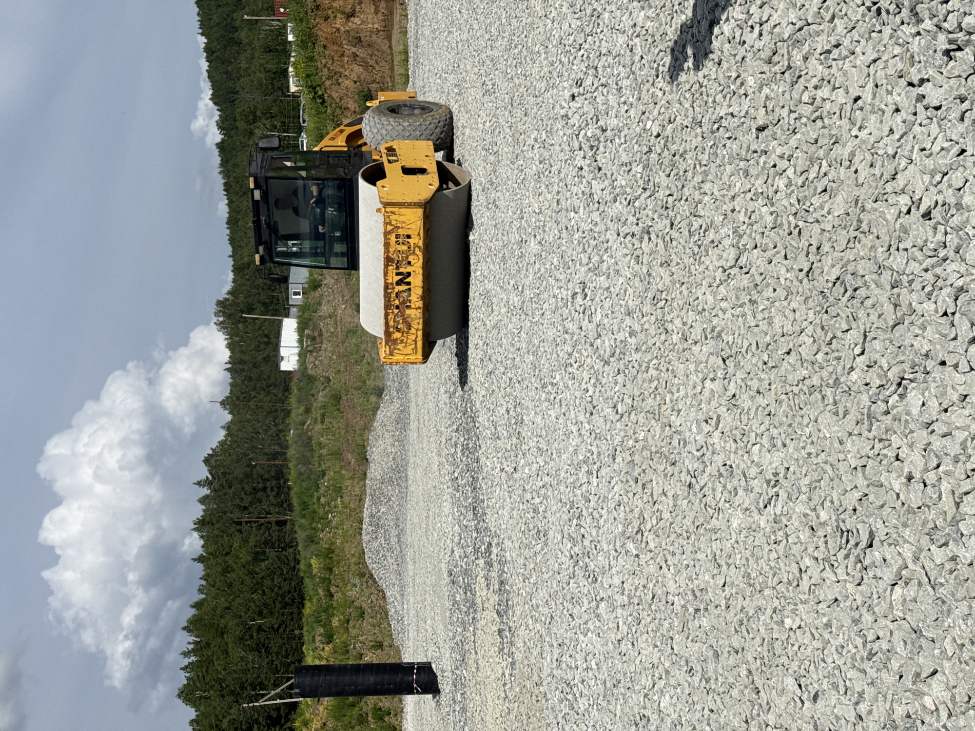 A yellow vibratory roller is compacting a surface covered in gray gravel. The roller has a cab with a person inside operating it. In the background, there is a pile of gravel, a dark cylindrical object, a line of trees, and several small buildings. The sky is cloudy. The image appears to have been taken in Leninsk, Russia.
