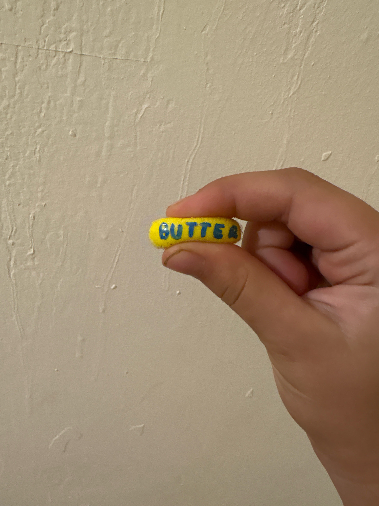 A close-up shot shows a hand holding a small, yellow, pill-shaped object with the word "BUTTER" written in blue letters on its side. The object is positioned in the center of the frame, with the hand gripping it from the right. The background is a textured, light-colored wall. The image does not contain information to determine the location in the city of Ashvem, India.