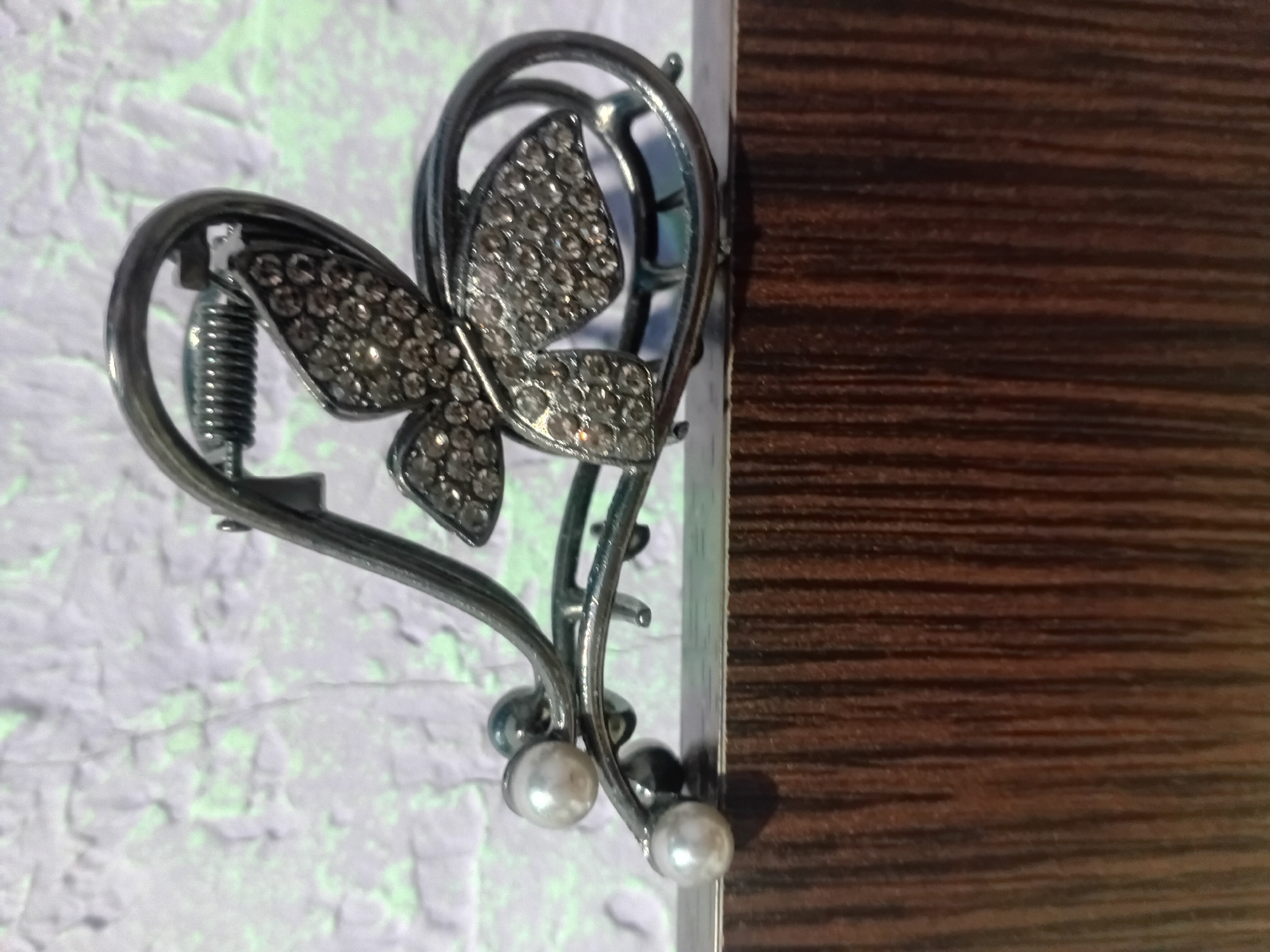 This media file features a close-up, eye-level shot of a decorative hair clip. The clip is shaped like a heart, with a butterfly design adorning its center. The butterfly's wings are encrusted with small, clear rhinestones, giving it a sparkling appearance. The heart shape itself is made of a dark, metallic material, possibly pewter or a dark-colored metal alloy. A coiled spring is visible within the heart's structure, suggesting the clip is functional. The clip is also embellished with two small, white pearl-like beads at the bottom.
The hair clip is positioned on the edge of a surface with a dark wood grain pattern, reminiscent of furniture or a door. The background is a textured, light-colored surface with a faint green tint, which appears to be a wall or fabric. The lighting suggests it is daytime, with the light source coming from the left, casting subtle shadows. The overall scene is simple and focuses on the intricate details of the hair accessory. There is no text visible in the image.