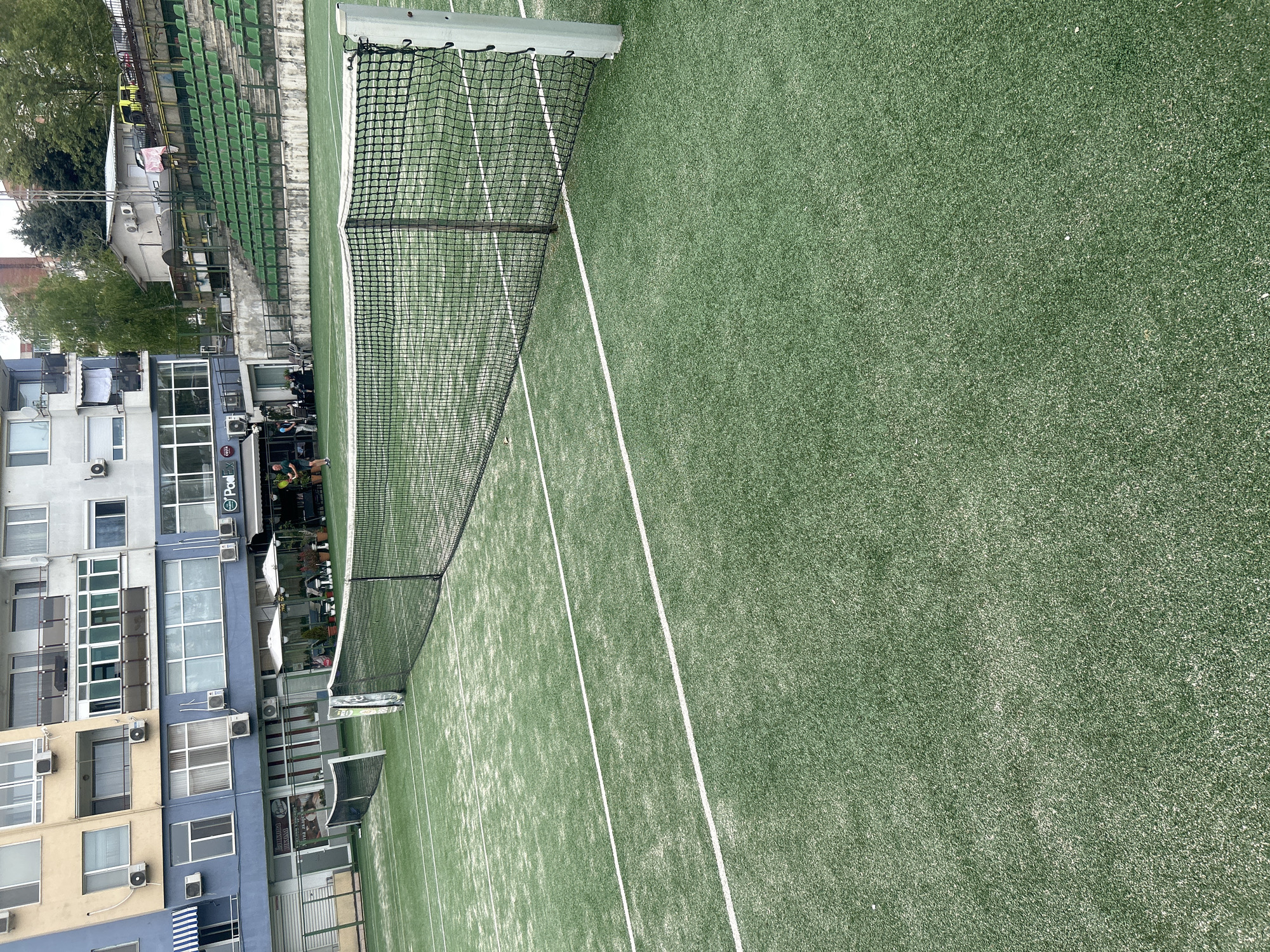 The image shows a tennis court with a green synthetic surface and white lines, a net in the middle. Adjacent to the tennis court is a multi-story building with blue and white facades and multiple windows. Green stadium-style seating is partially visible behind the tennis court. There are people visible at the end of the court. Given the location in Veliko Tarnovo, Bulgaria, this is most likely a padel or tennis court situated in an urban environment, possibly associated with a sports club or residential complex.