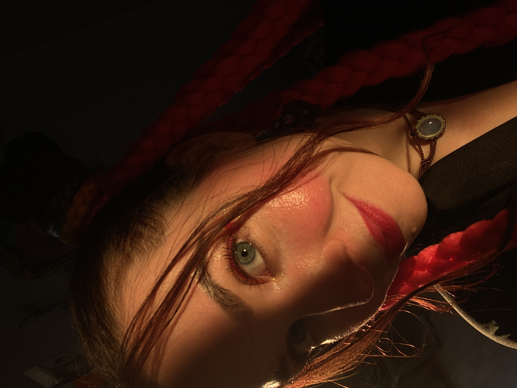 The image is a close-up, dimly lit portrait of a young woman with striking blue eyes and red lipstick. She has long, dark hair styled in braids with red extensions. Her face is illuminated by a strong light source, casting dramatic shadows. She wears a dark top and a necklace with a light blue stone. The background is dark and indistinct. There is no indication of the city of Chaikovsky, Russia, within the image.
