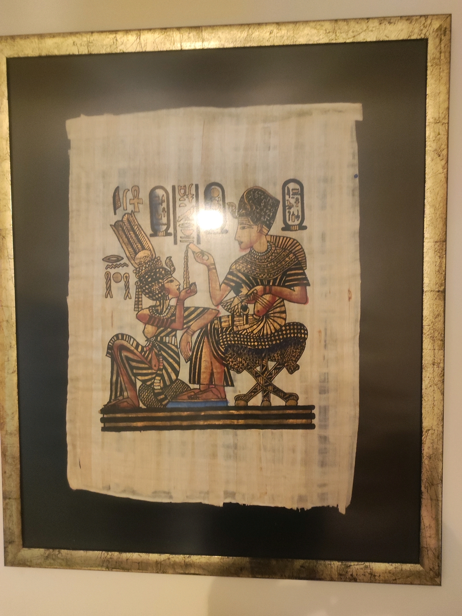 The image displays a framed Egyptian papyrus artwork. The artwork depicts two figures in traditional Egyptian attire. The figure on the left is kneeling and wearing an elaborate headdress and a striped garment. The figure on the right is seated on a stool, also elaborately dressed, and appears to be holding a string or object. Hieroglyphics are visible above and to the side of the figures. The papyrus is mounted against a dark background and enclosed in a decorative gold frame. The photograph is taken from a slightly upward angle, showing the framed artwork hanging on a wall. The location within Khazar Raion, Azerbaijan is not discernible from the image itself, as the image solely focuses on the artwork and its presentation.