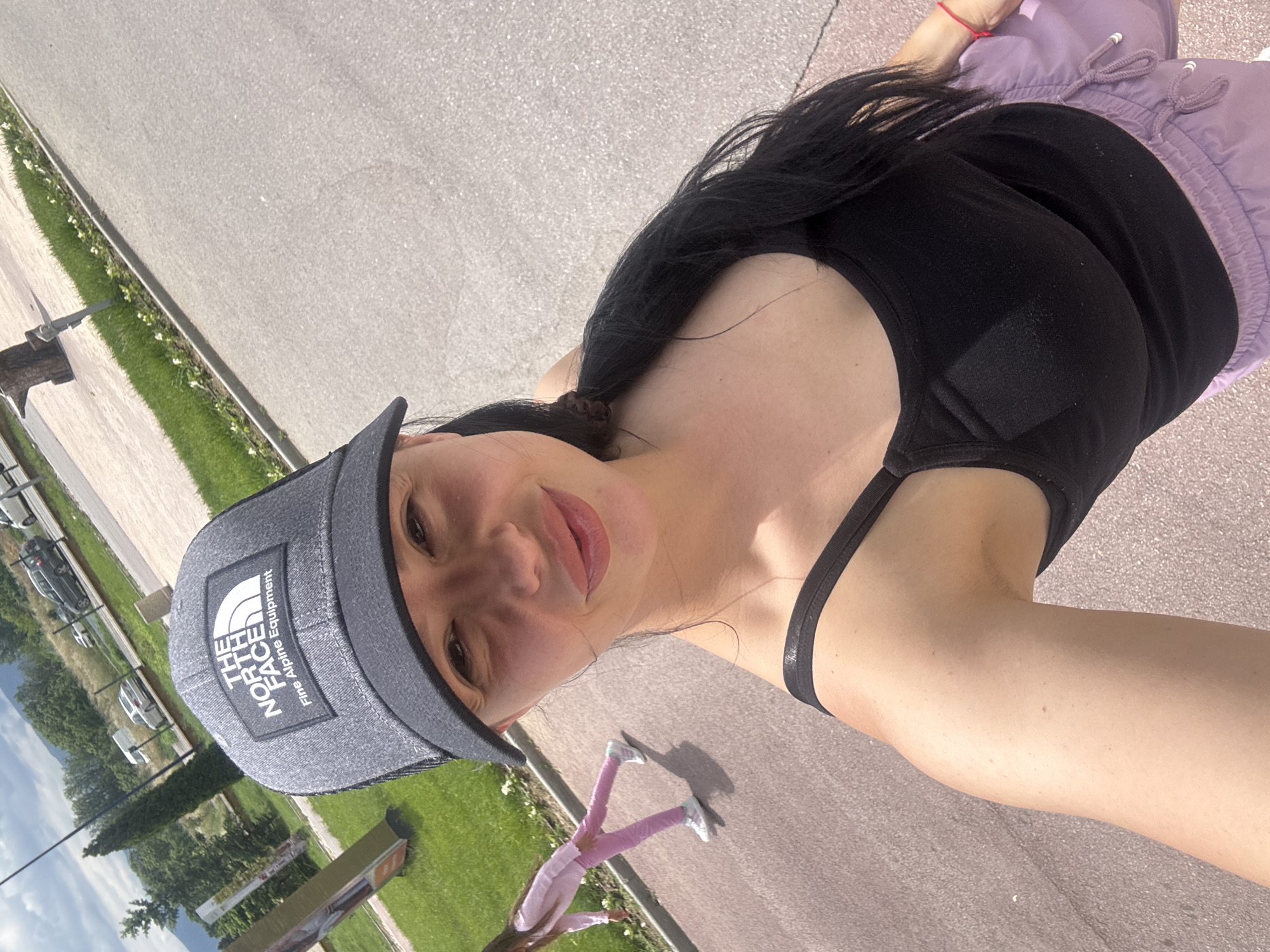 A woman with long, dark hair pulled back into a ponytail, wearing a black tank top, light purple shorts, and a gray The North Face baseball cap, is taking a selfie outdoors. She is standing on a paved surface next to a strip of green grass and white flowers. In the background, there is a grassy area with trees and parked cars. A small child wearing pink clothing and white sneakers is visible near the bottom of the frame. The scene appears to be a sunny day.