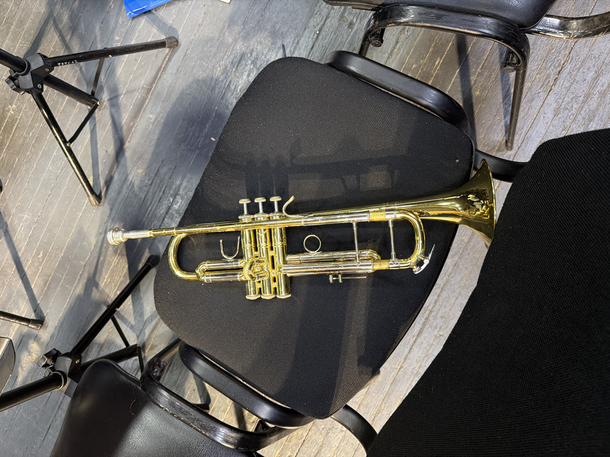 The image captures an overhead view of a brass trumpet resting horizontally on a black fabric chair. The trumpet is primarily golden-colored with silver valve tops and a silver mouthpiece, displaying its intricate tubing and three valves.
The chair supporting the trumpet has a black fabric seat and back, and a black metal frame. Surrounding it, several other chairs are partially visible; some match the fabric chair, while others appear to have worn black faux leather seats and backs, also with black metal frames.
To the left of the central chair, a black metal music stand with a tripod base is positioned on a worn wooden floor. The floor consists of greyish-white painted or stained planks, showing visible signs of use.
No individuals are present in the scene, which suggests a quiet moment, possibly before or after a rehearsal or performance. The setting, with multiple chairs and a music stand, points towards a music practice room, classroom, or a stage area.
On one of the black metal legs of the music stand, a faint white marking or label is visible, which appears to read "TRV_42". The lighting is consistent with an indoor environment, and no specific time of day or outdoor conditions can be determined.
