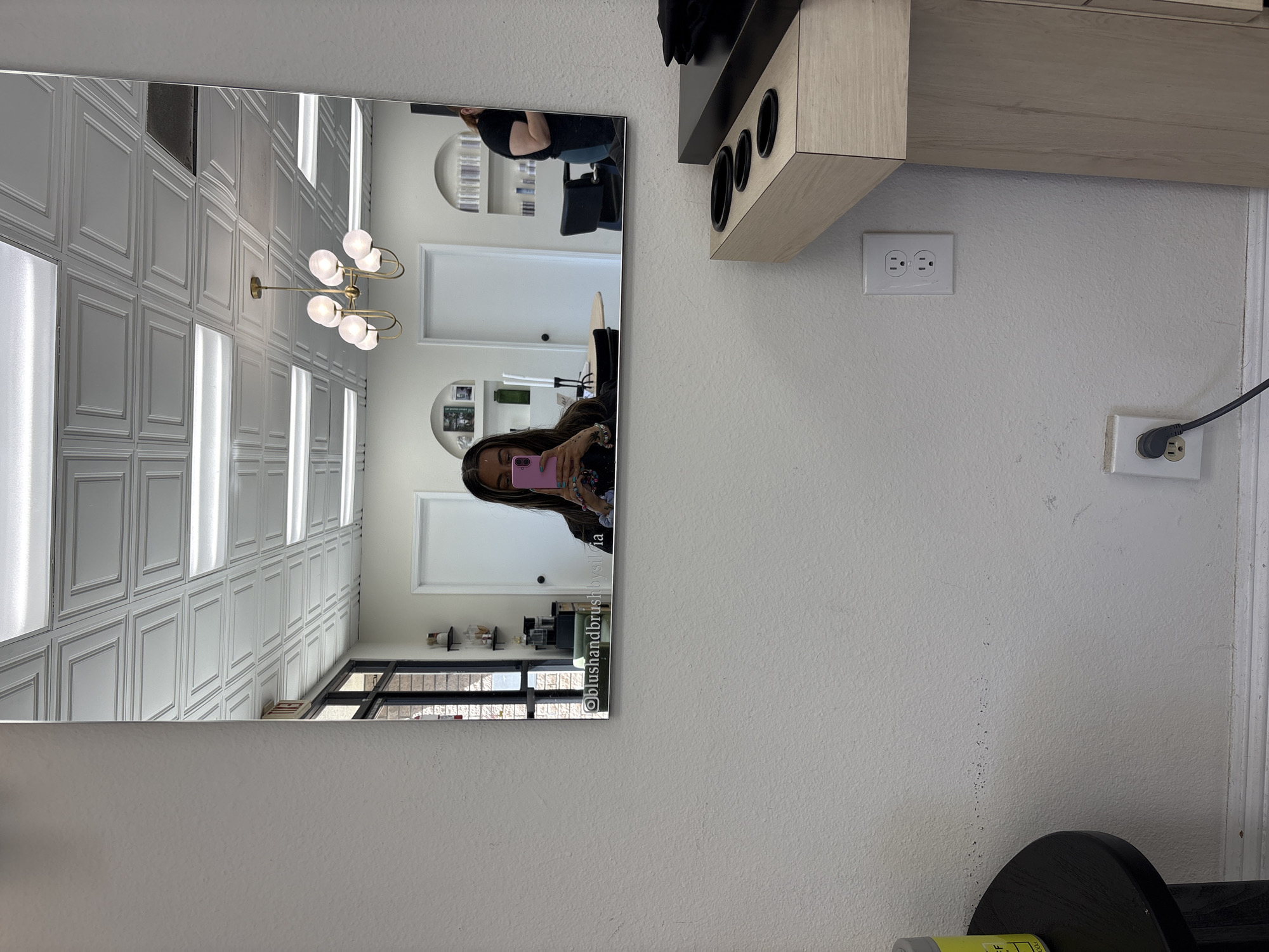 The image displays a selfie taken in a large, rectangular mirror, reflecting a modern hair salon interior. A person with long dark hair, wearing a dark top, is seen holding a pink smartphone to capture the image, with their face partially obscured by the device. To their left in the reflection, another individual, possibly a stylist, is partially visible in a black shirt, positioned near a black salon chair.
The salon's interior, as depicted in the mirror's reflection, features white paneled walls and a ceiling adorned with bright, linear strip lighting and a decorative brass-colored chandelier with multiple spherical, illuminated bulbs. Additional elements in the reflection include salon chairs, shelves displaying various hair products, white doors, and a black-framed window.
Outside the mirror, on the textured white wall, a light wood-colored shelf with three black circular inserts (likely for holding hair tools) is mounted. Below the shelf, two white electrical outlets are visible, with a black power cord plugged into the lower one, extending out of frame. The white wall exhibits some minor scuff marks and dirt specks.
Visible text on the lower right corner of the mirror's surface reads "@blushandbrushbysilvia," indicating the salon's social media handle. The scene portrays an indoor setting, likely during the day, within a clean and well-lit hair salon in Bellflower, United States of America.