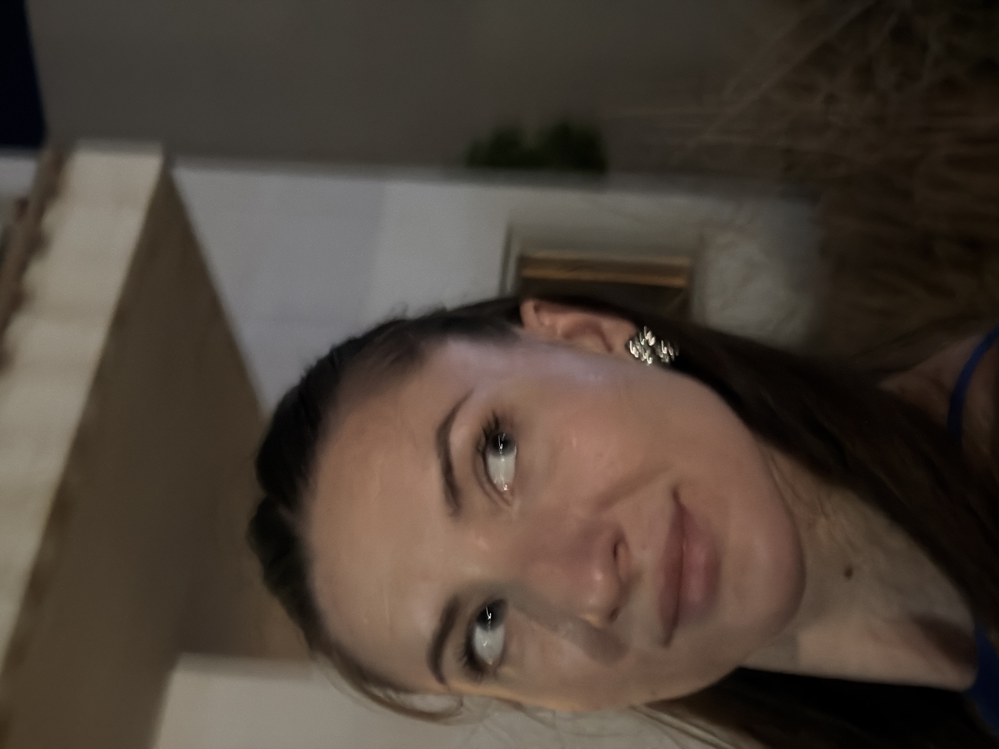 A woman is captured in a close-up, medium-shot photograph, positioned on the left side of the frame, looking to her left with her eyes wide. She has long, dark brown hair, with some strands pulled back from her face, and she is wearing a silver, floral-shaped earring on her right ear. Her skin appears smooth, with subtle makeup highlighting her features. She is wearing a blue garment, visible at the bottom of the frame. The background is softly blurred, suggesting an indoor or nighttime setting. The architectural elements hint at a location in Sharm El Sheikh, Egypt, possibly a resort or a public space with ambient lighting. The overall lighting is dim, with warmer tones in the background and more neutral lighting on the woman's face.