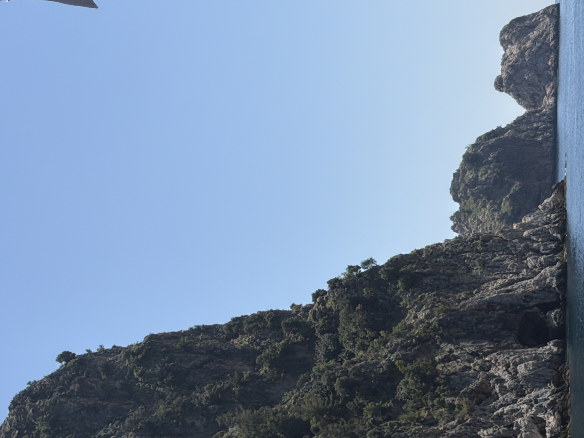 The image captures a rugged, rocky coastline in Alanya, Turkey. The steep, mountainous terrain is covered in sparse vegetation. The right edge of the frame shows a body of water, presumably the Mediterranean Sea. The sky above is clear and light blue.
