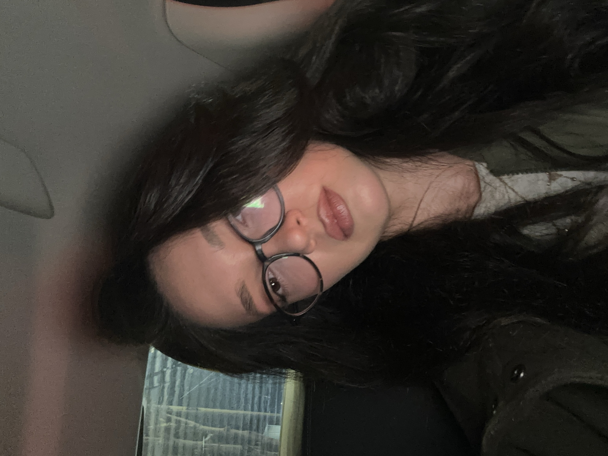 A female individual is depicted in a close-up, head-and-shoulders portrait, positioned within what appears to be the interior of a vehicle. She has long, dark, wavy hair, and wears dark-rimmed eyeglasses that show reflections of an unseen light source. Her face exhibits light-toned skin, subtle eye makeup, and full lips enhanced with a light-colored, glossy product. She is dressed in a dark olive-green jacket over a lighter, possibly patterned, top, with a thin necklace visible. Her gaze is directed straight into the camera, maintaining a neutral expression. The background on the left side shows a blurry, indistinct view through a window, revealing faint horizontal elements, possibly industrial structures or blinds, in a dimly lit environment. The upper portion of the image shows the grey interior roof lining of the vehicle. No specific landmarks or distinguishing features are present to identify a precise location within the city of Yerevan, Armenia.