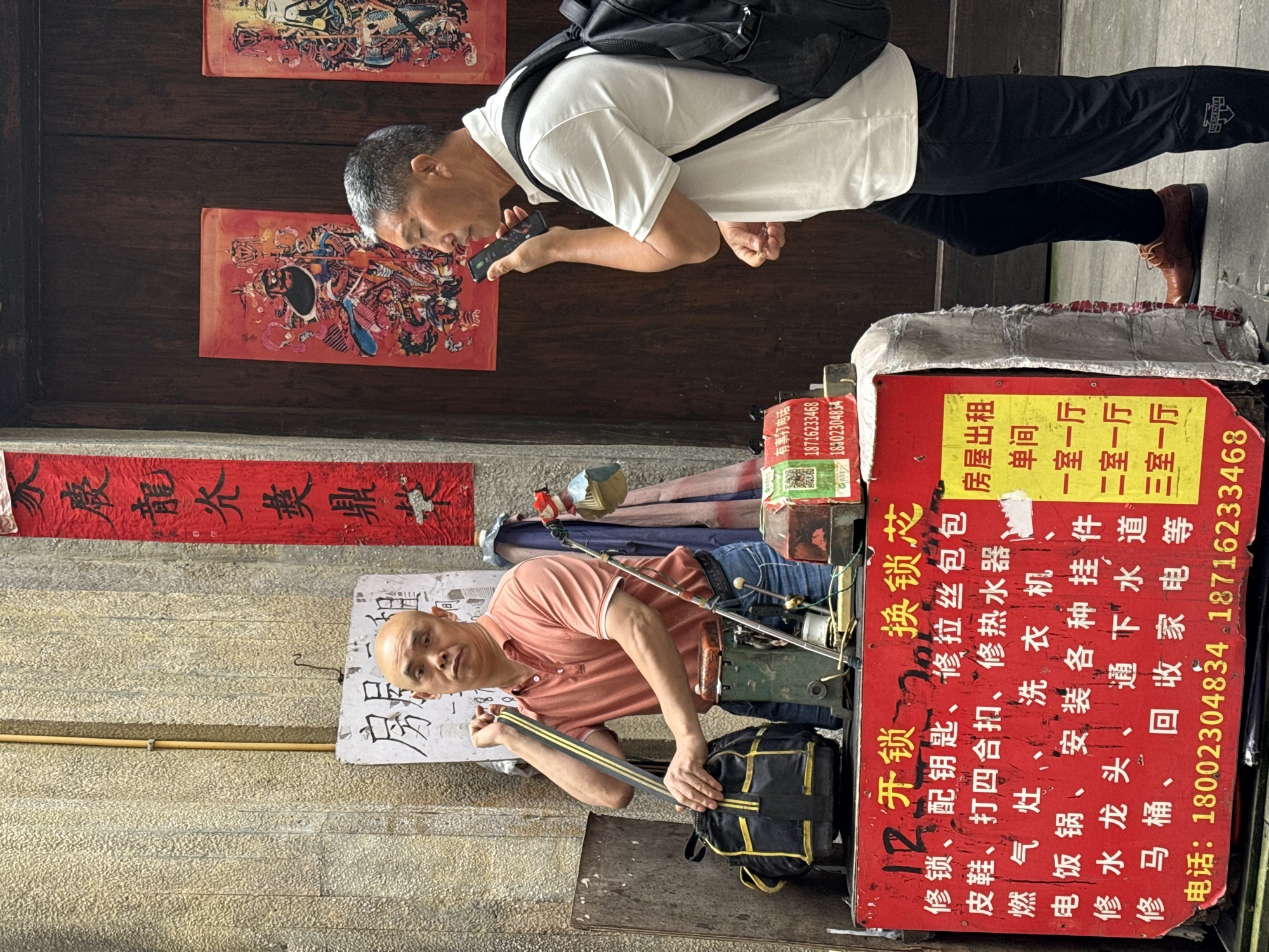 Here is a description of the image:
The photo depicts a scene from Yuzhong District, China, likely along a narrow street or alleyway. Two men are visible, one standing and the other seated. The standing man wears a white t-shirt, black pants, brown shoes, and a backpack. He is looking down at his phone, which he holds in his hand. The seated man wears a peach-colored polo shirt. He sits behind a small, portable workstation or stand, and he is holding a tool in his hands. The stand is cluttered with various tools and items related to his trade, which includes locksmithing, shoe repair, and appliance repair. A red sign on the stand lists the services offered and includes phone numbers. Calligraphic banners adorn the wall behind the stand. Additionally, images featuring Chinese figures hang above the man looking at his phone. The environment appears to be a traditional urban area, with stone or brick walls and narrow passages.