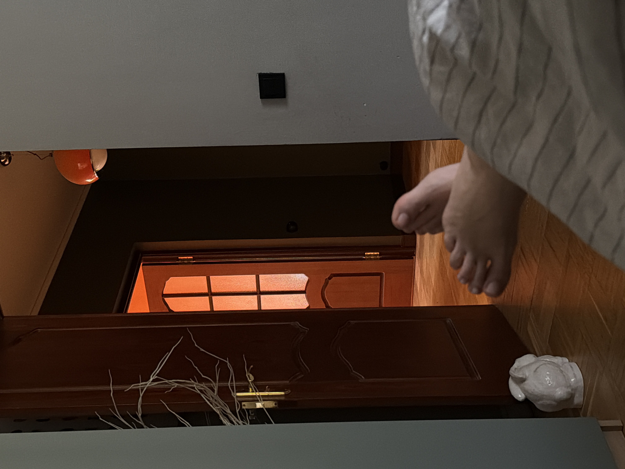This image depicts an interior scene in Khimki, Russia, from a low-angle perspective, likely from a bed. In the immediate foreground, a person's bare feet are visible, extending from beneath a light-colored duvet or blanket with thin, dark horizontal stripes. To the left, a portion of a light grey wall or furniture panel is visible, next to which a dark brown wooden door with a brass handle is slightly ajar. A decorative arrangement of white, dry-looking branches is positioned near the door's handle. On the light brown parquet flooring below, a white ceramic rabbit figurine rests. To the right, a segment of a light grey wall features a black square light switch. Through the partially open wooden door, a second room is visible, characterized by a strong orange-red glow emanating from within, illuminating another dark brown wooden door with an arched top and multiple glass panels arranged in a grid pattern. This inner door is also slightly ajar, revealing a darker space beyond it. A portion of an orange and white spherical ceiling light fixture is visible in the upper right background. The overall lighting presents a contrast between the dimmer, natural light in the foreground and the intense orange illumination from the adjacent room.