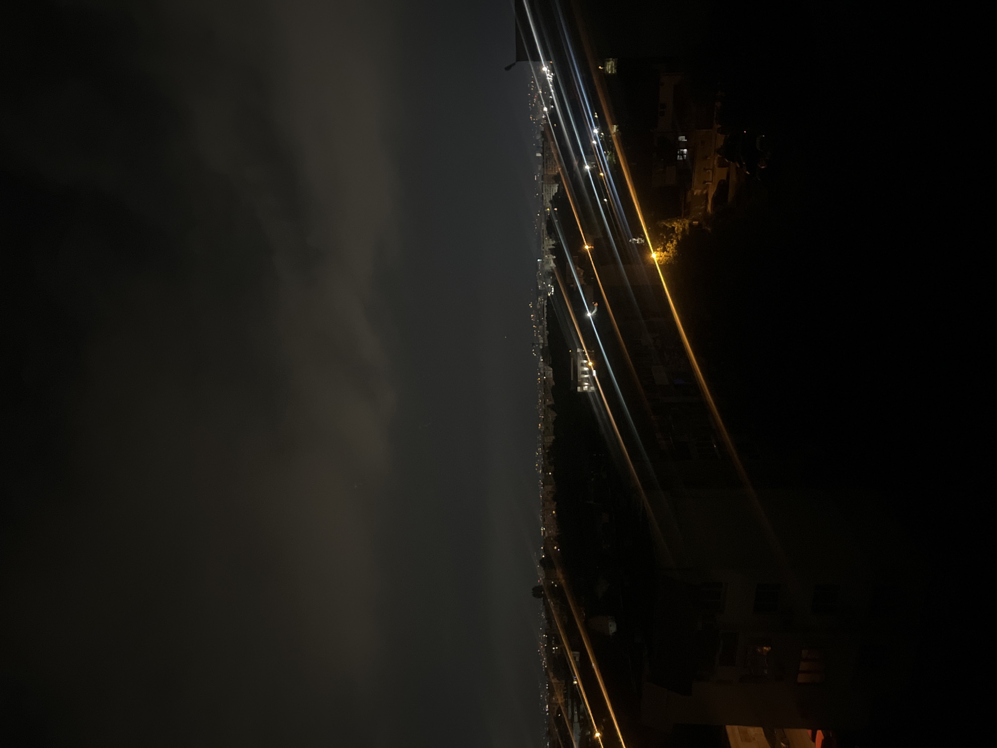 The image shows a night view of Sofia, Bulgaria, taken from an elevated position. The left side of the image is dominated by dark clouds, while the right side features a city skyline with buildings and streetlights. Several bright streaks of light traverse the scene, possibly from passing vehicles or camera movement. The image is dark, with only the city lights providing illumination. The photograph appears to have been taken from a high-rise building.