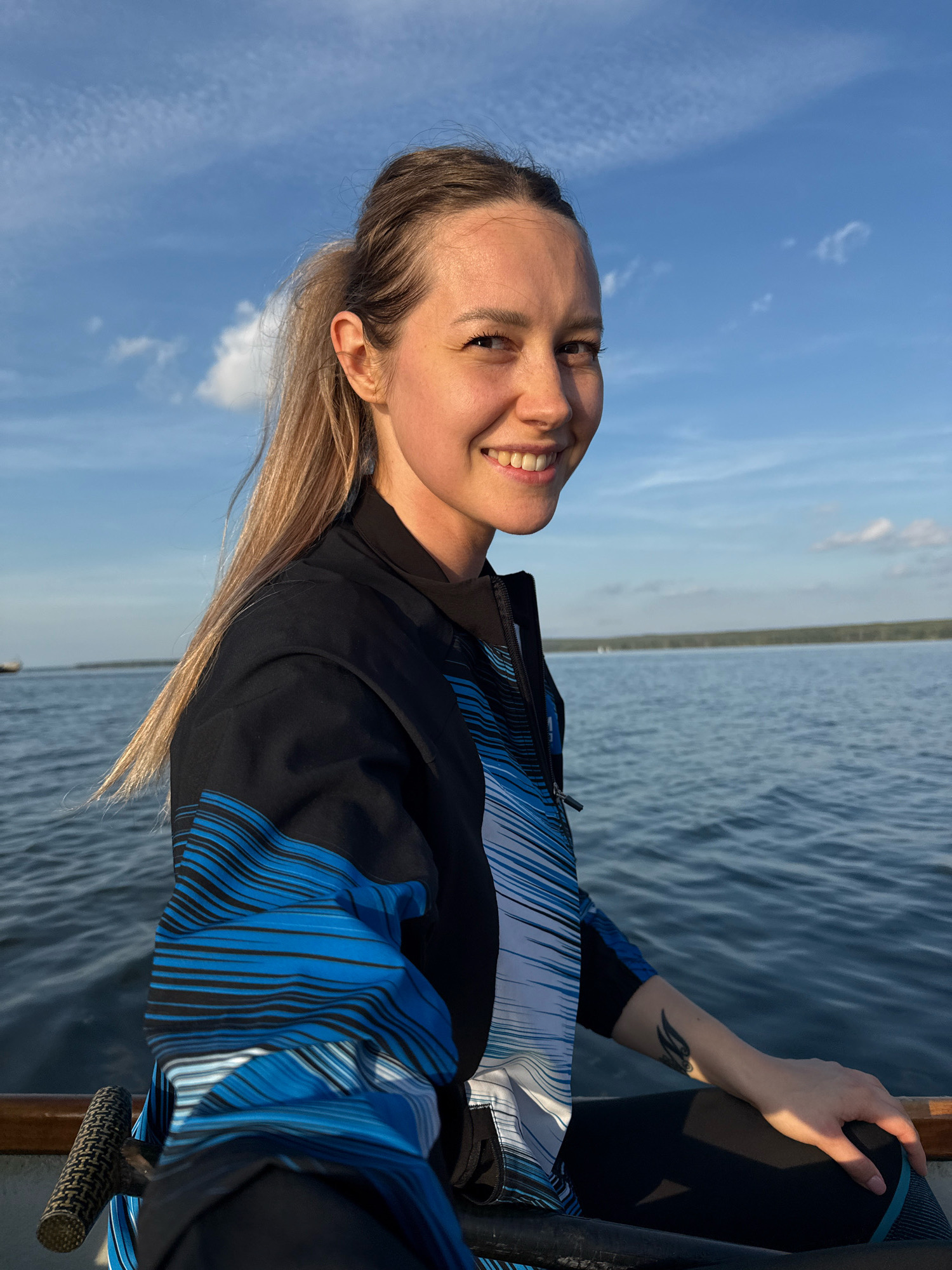 A young woman with blonde hair in a ponytail is looking over her shoulder and smiling at the camera. She is wearing a black jacket with blue and white striped detailing. She is sitting in a boat on a body of water, with the sky and clouds visible in the background. The location is likely near Chaikovsky, Russia, as indicated by the presence of the Kama River, a prominent feature of the city's geography. The overall scene suggests a leisurely activity on the water.