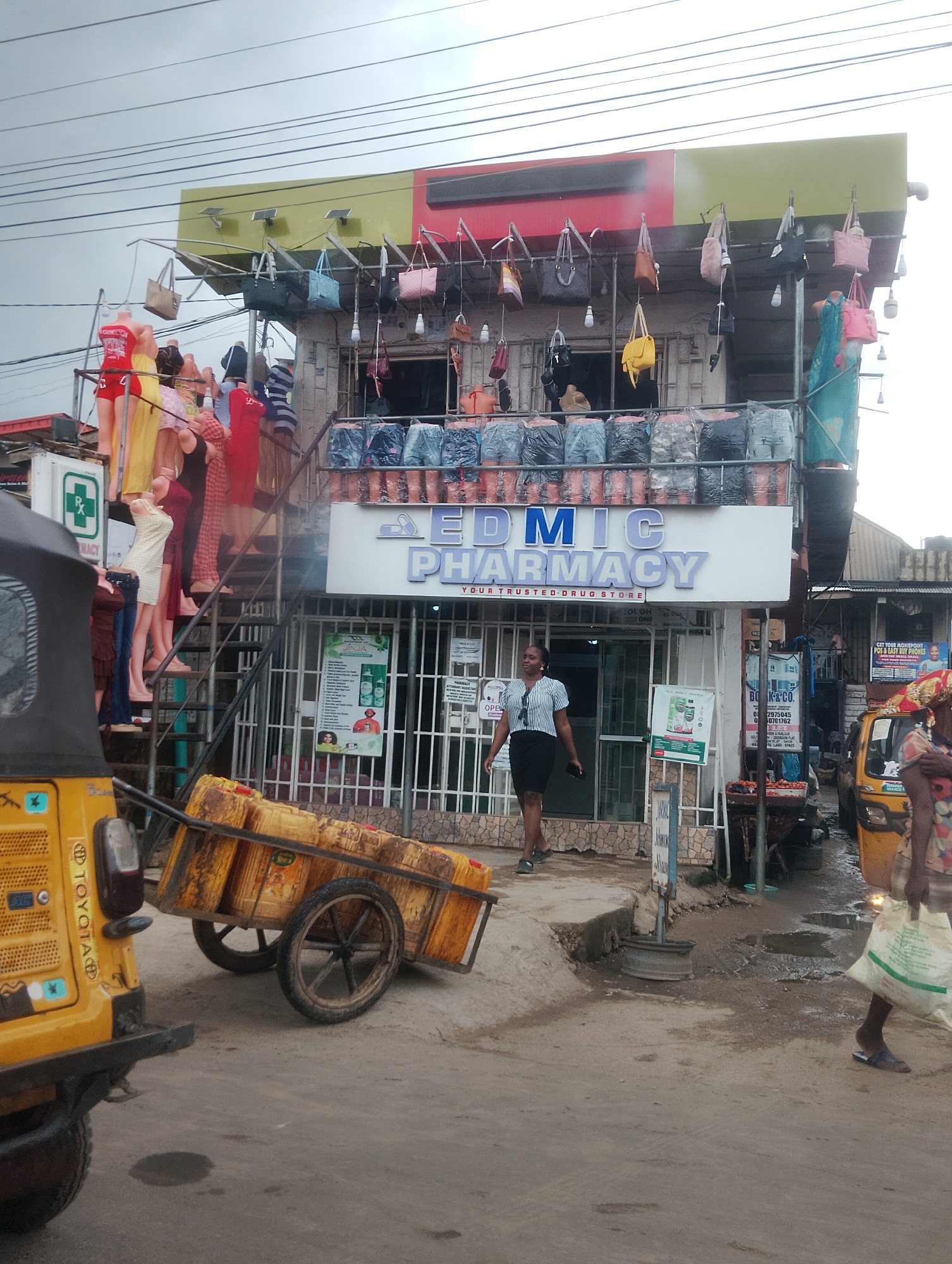 This is a full-shot, eye-level, outdoor photograph taken on a cloudy day in Iju, Nigeria. The scene depicts a street view of a business establishment with a pharmacy sign that reads "EDMIC PHARMACY YOUR TRUSTED DRUG STORE." The building has multiple levels, with various items displayed for sale. On the upper level, mannequins are dressed in clothing, and handbags are hung from the storefront. The lower level features storefront windows, and a woman in a striped shirt and dark skirt is walking past the entrance. To the left of the building, a yellow tricycle or auto-rickshaw is parked, and a cart filled with yellow jerrycans is being pushed. In the foreground, the street is paved with asphalt and shows signs of recent rainfall, with puddles of water visible. On the right side of the image, another woman is walking with a white bag. The overall environment suggests a bustling marketplace or commercial area.