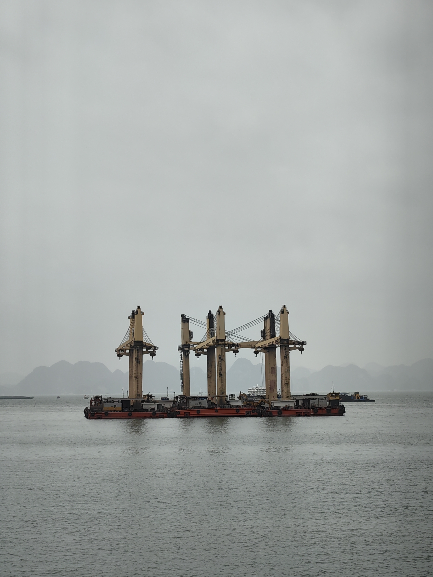 The image depicts a barge in Hong Gai Ward, Vietnam, with four large port cranes mounted on it. Mountains are visible in the background, obscured by a cloudy sky. The water is choppy. Other boats are in the distance.