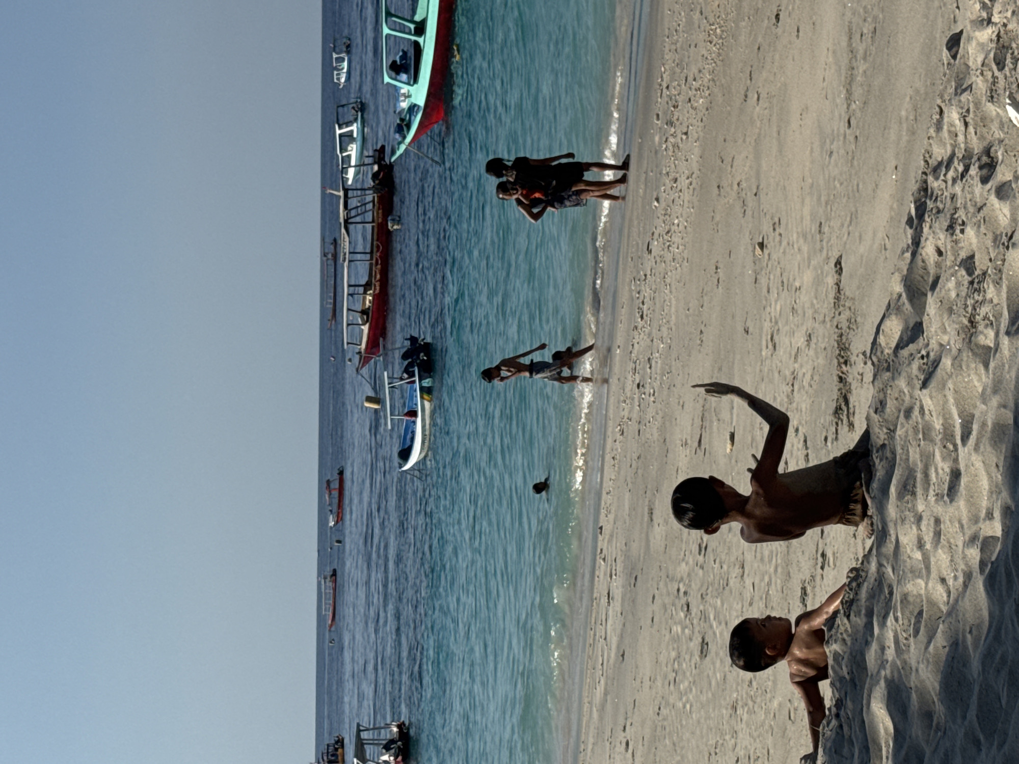 This media file depicts a sunny day at a beach in an unknown Indonesian location. The scene features several wooden boats docked in the shallow, clear blue water. Some of the boats are colorful, with one prominent green and red vessel and another dark red boat. Further out in the water, a person is swimming.
On the sandy shore, there are several children. Two young boys are the closest subjects, with one leaning against a rocky outcropping and the other standing and looking out with an arm raised. Further up the beach, a group of three children are standing together. One of the children appears to be playing with or interacting with another person. The children are all wearing casual clothing.
The bright sunlight suggests it is likely midday or early afternoon. The weather appears to be clear and warm, ideal for a beach outing. The presence of multiple boats and the children playing in the water and on the sand indicate a relaxed and leisurely atmosphere. There is no visible text in the image.