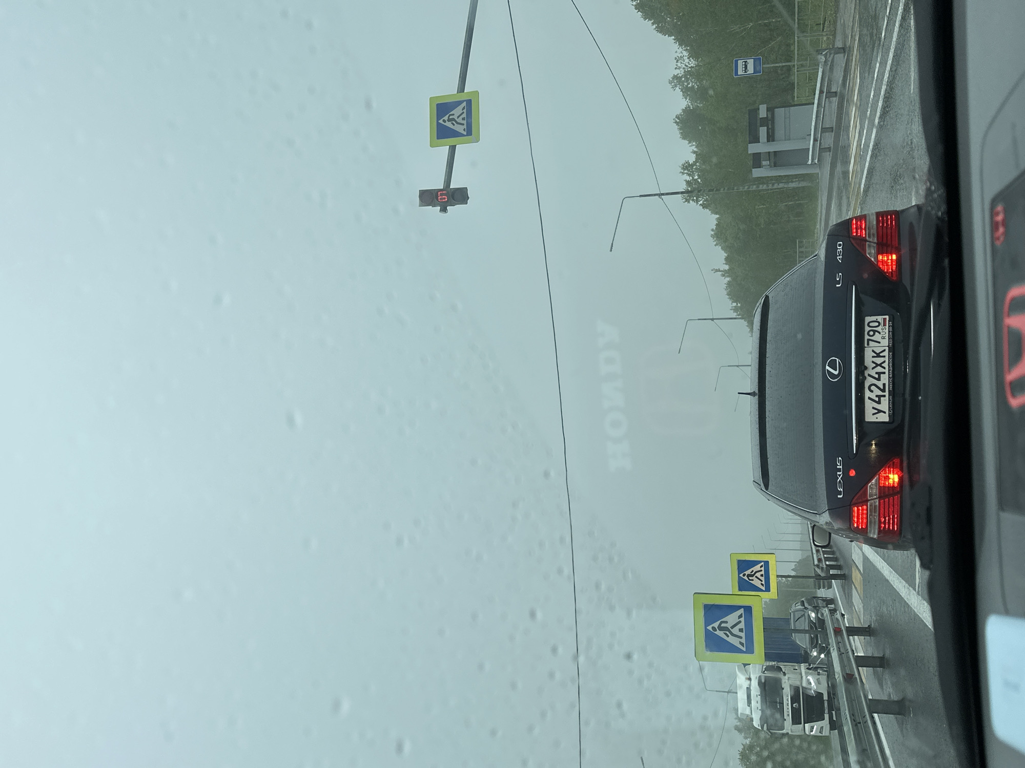 The image depicts a rainy day on a multi-lane road in Pionersky, Russia. The perspective is from inside a vehicle, looking forward. A black Lexus LS 430 is in the foreground, with its rear lights illuminated. The road surface is wet, indicating recent rainfall, and water droplets are visible on the windshield.
Several traffic signs are present. Two pedestrian crossing signs, one larger than the other, are visible on the right side of the road. Further ahead, a traffic light with a red display showing "07" is illuminated.
In the distance, a white truck is on the left lane. The surrounding environment features dense green trees and a grey, overcast sky. The overall atmosphere suggests a traffic situation during inclement weather.