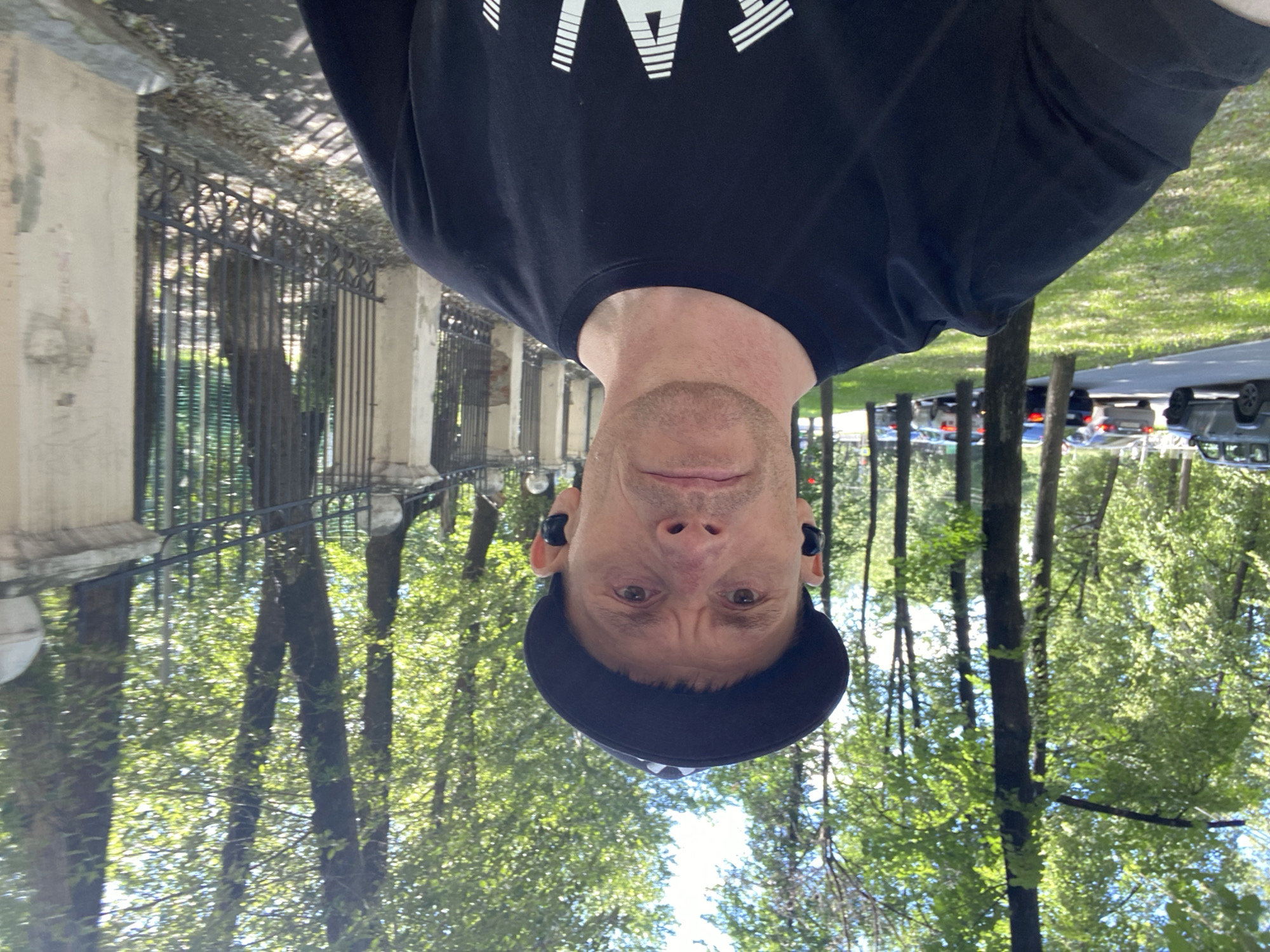 In an outdoor, daytime scene in Yekaterinburg, Russia, a light-skinned man wearing a black baseball cap and a black t-shirt with white text on it is captured in a selfie. He has short, light brown hair and is wearing black earbuds. The man's expression is neutral as he looks directly at the camera. He is positioned in the foreground, with a blurred background that includes trees, a black metal fence, and a light-colored wall. To the left of the man, a paved path and a grassy area are visible, with several cars parked along the road. The sunlight suggests a clear day.