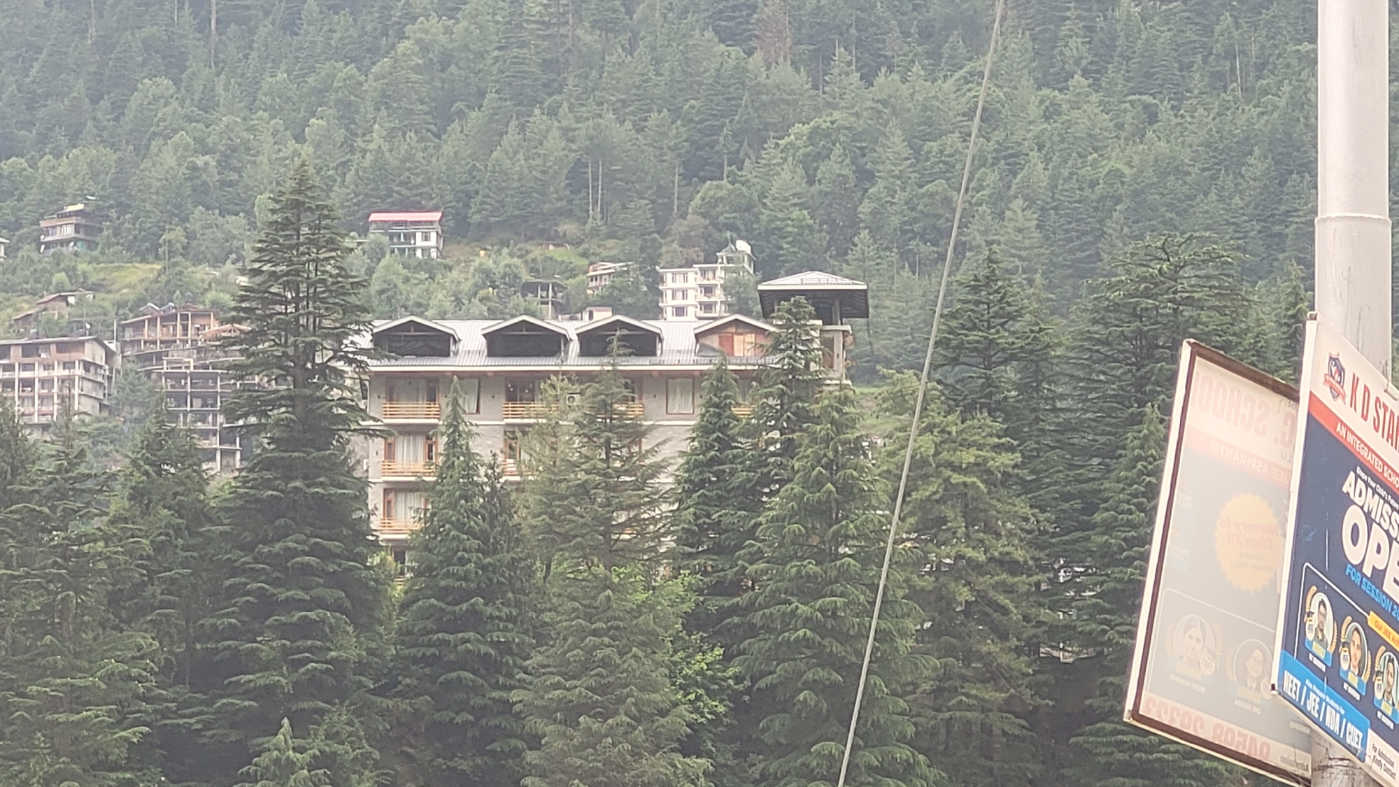 The image captures a view of buildings nestled on a hillside covered in dense pine forests, likely in Vashisht, India, known for its mountainous terrain and guesthouses. A multi-story hotel or guesthouse, characterized by its balconies and dark sloped roofs, is prominently featured in the foreground. Other buildings, some appearing under construction with exposed concrete frameworks, are visible further up the slope. The environment is dominated by evergreen trees, suggesting a mountainous region. In the lower right corner, a portion of a white pole is visible, with several advertisements or signs attached to it. One sign displays the text "KD STAR" and "ADMISSION OPEN," indicating a school or educational institution. The overall scene suggests a daytime view, with overcast or hazy conditions contributing to a subdued lighting.