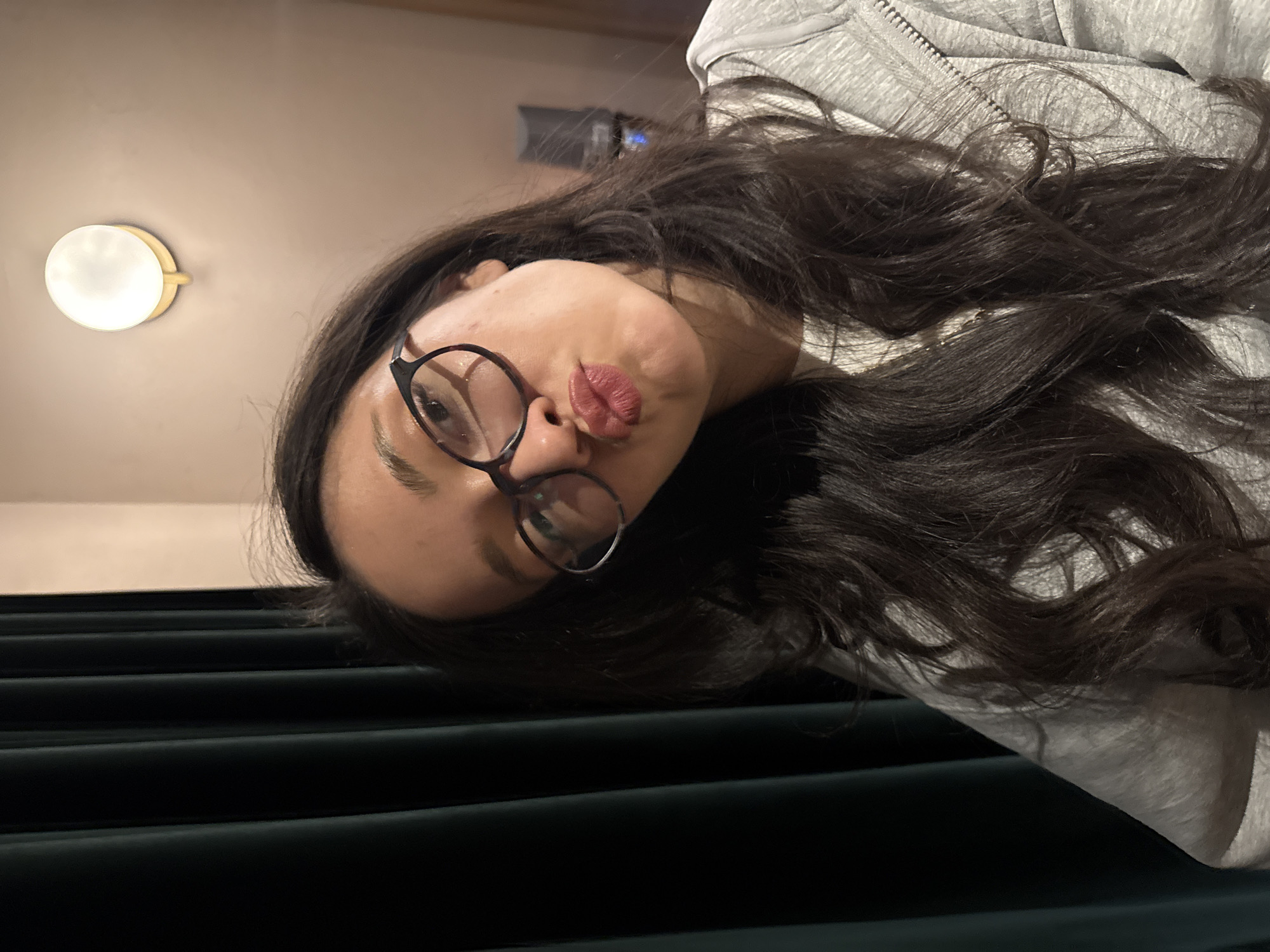 A selfie captures a person with long, dark brown hair and glasses, positioned at an unusual angle against a dark green, plush-looking horizontal surface, possibly a cushioned bench or seat. The person has their lips pursed in a kissing gesture. They are wearing a light grey hooded sweatshirt. In the background, a wall sconce emits a warm light. Based on visual cues and the prompt's request, the location is Yerevan, Armenia.