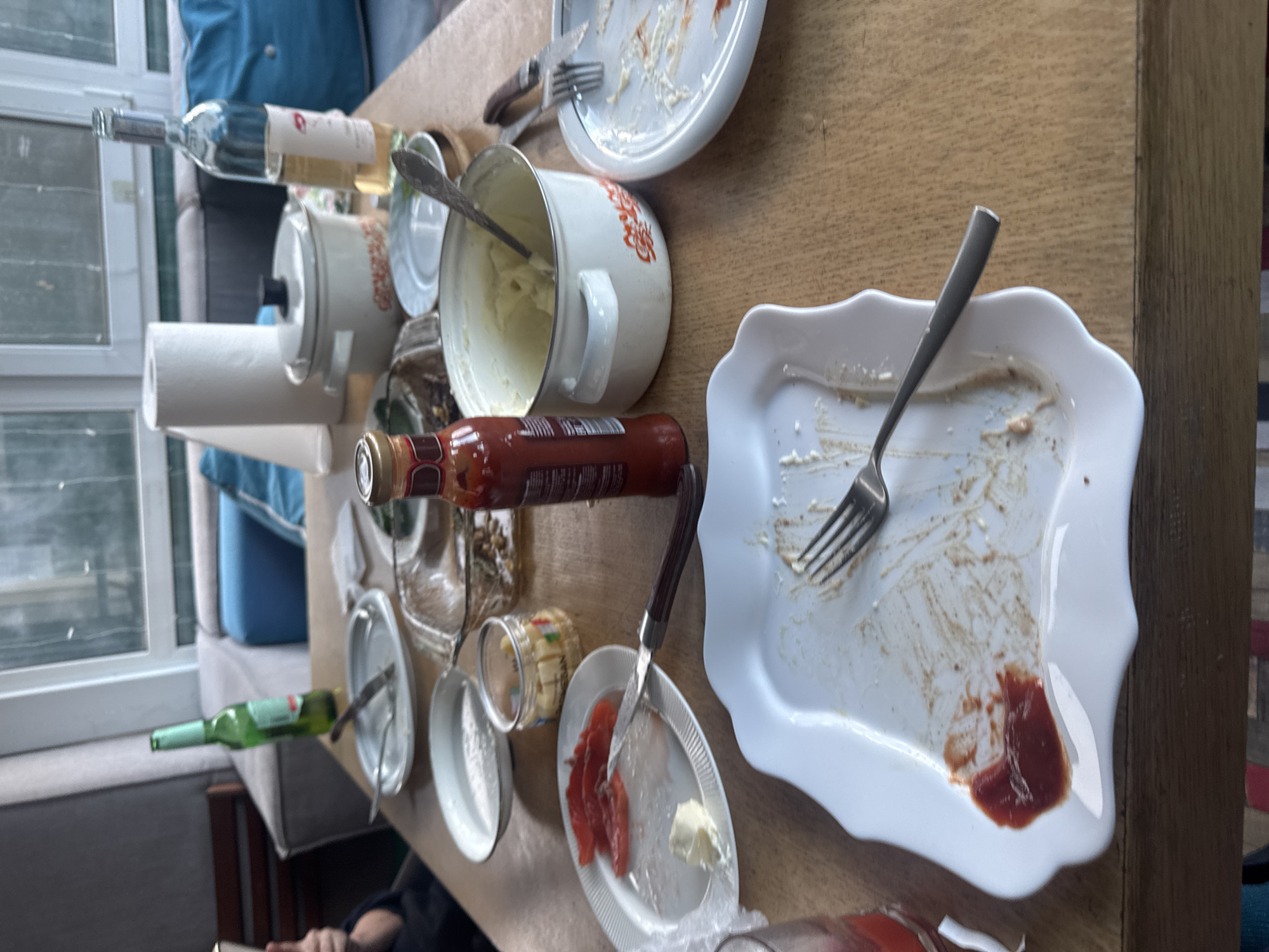 The scene depicts a dining table in Ptichnoye, Russia, covered with the remnants of a meal. Several empty plates, some with food residue, are visible, alongside used silverware, including forks, knives, and spoons. A white enamel pot containing mashed potatoes sits near a partially full bottle of ketchup. Other containers hold unidentified food items. Bottles of wine and a paper towel dispenser are also present. A window is visible in the background, and a sofa with cushions. The focus is on the disordered state of the table after a meal, with no people clearly visible, only a partial view of someone's clothing in the lower left.