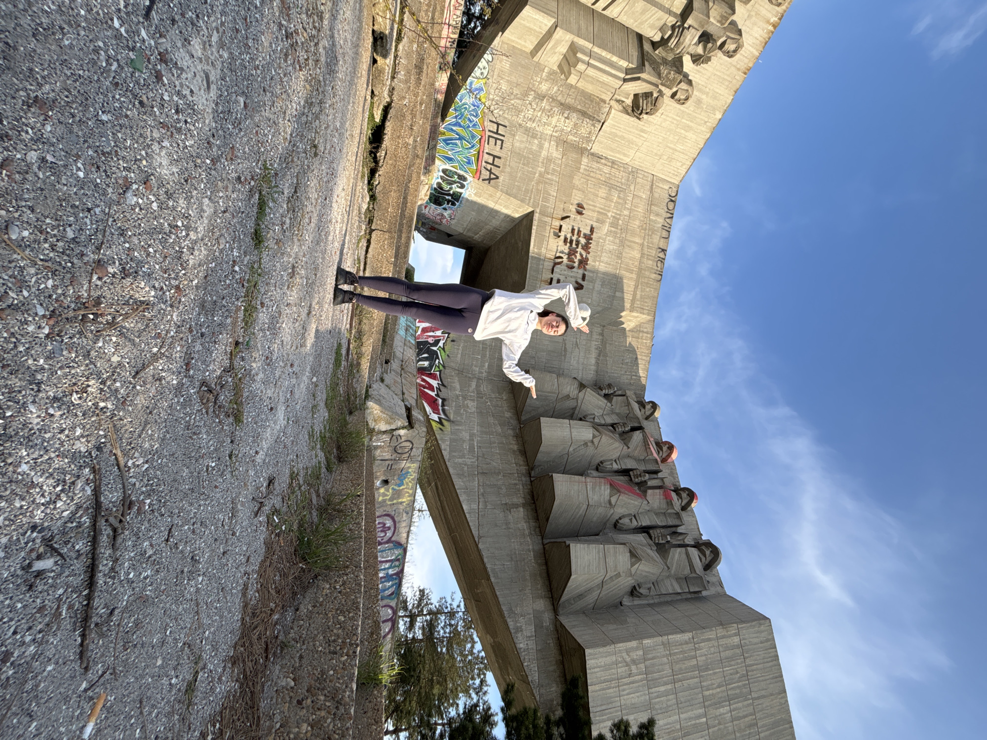 The image shows a person standing in front of a large concrete monument, possibly in an urban setting. The monument features sculpted figures and is covered in graffiti. The person is wearing a white jacket and dark pants. The sky is blue with some clouds. The location appears to be an abandoned or neglected area within the city.