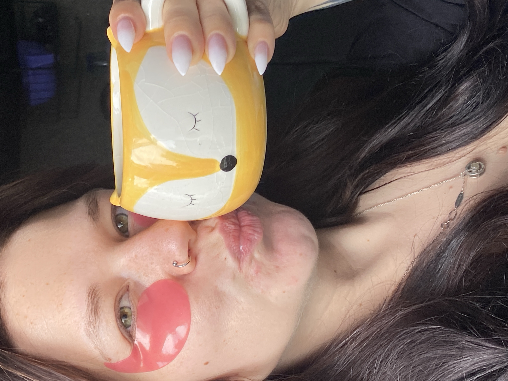 A close-up, upward-angled shot captures a person, likely a woman, playfully holding a fox-shaped mug to their face. The person's dark hair cascades around their head, suggesting they are lying down. Their left eye, with a green iris, is visible, along with a silver hoop nose ring on their right nostril. Pink, gel-like under-eye patches are applied beneath their left eye, indicating a self-care or relaxation routine. Their lips are pursed in a puckered expression, making contact with the base of the yellow and white fox mug.
The ceramic mug is designed with a stylized fox face, featuring closed black eyes and a black nose. The person's hand, adorned with pointed, white-tipped ombre nails, grasps the mug. A delicate silver necklace with small charms is visible around their neck. The background is dark and out of focus, with a subtle blue glow on the left, possibly a reflection from a screen. The image conveys a lighthearted and relaxed mood, set indoors in Chaikovsky, Russia. No visible text appears in the image.