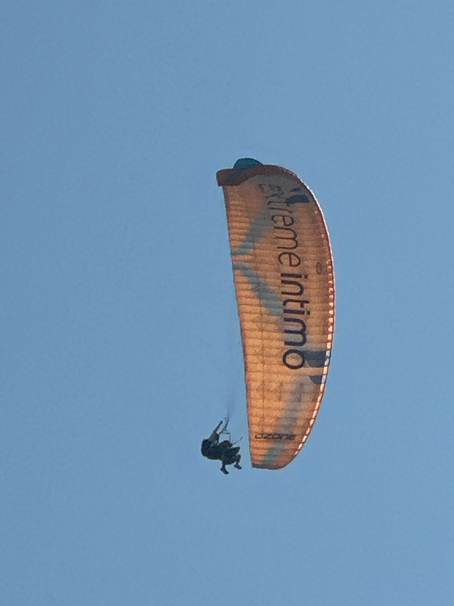An orange and blue paraglider with the words "xtreme Intimo" is soaring through a clear blue sky, with a person in a dark harness suspended beneath it. The paraglider is partially silhouetted against the sky, and the person appears to be in a relaxed flying posture. The image appears to have been taken in Boreti, Montenegro, although there are no other discernible landmarks to confirm this location. The overall impression is one of freedom and adventure.