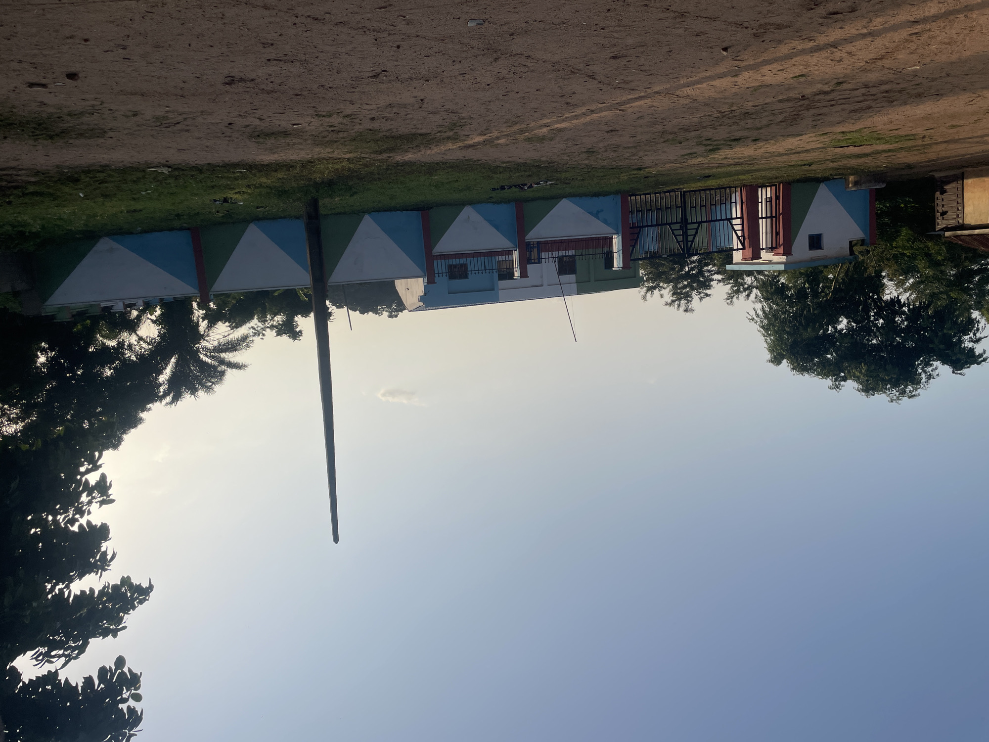 This media file depicts a scene in Kabba, Nigeria, likely in the late afternoon or early evening, given the soft, diffused light. The primary subjects are a series of buildings, possibly a school or community center, characterized by a colorful, geometric facade. The walls are adorned with a pattern of white, blue, and green triangles, creating a vibrant and distinctive appearance. Behind these buildings, a black metal gate is visible, suggesting an entrance or boundary.
The setting appears to be an outdoor area with a dirt or gravel ground surface in the foreground and a strip of green grass along the base of the buildings. Lush green trees, including a palm tree, frame the scene, some silhouetted against the bright sky. A tall, dark pole stands prominently in the mid-ground, its purpose unclear from the image alone. The sky is clear and bright, indicating fair weather.
There are no people clearly visible in the image, nor any specific activity taking place. The focus is on the architecture and the surrounding environment. The overall impression is one of a well-maintained, possibly institutional or public space in a tropical or sub-tropical location. The bright colors of the building's facade might suggest a cheerful or welcoming atmosphere. No text is visible in the image.